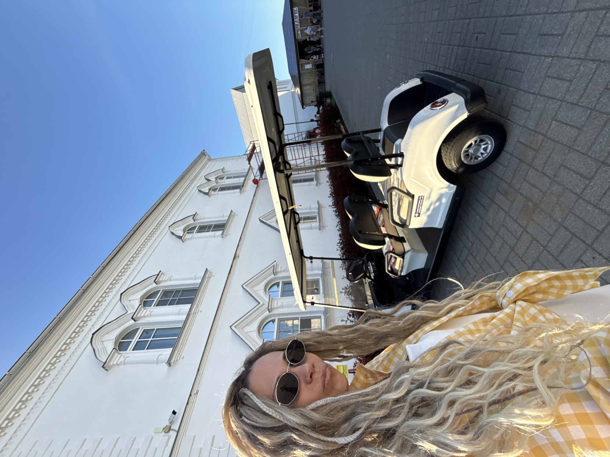 A female individual is positioned in the foreground, angled slightly left, with long, blonde and white wavy hair, interspersed with dreadlocks or braids. She wears dark, oval-shaped sunglasses and a yellow and white gingham (checkered) button-up shirt, tied at the front, over a white garment. Her lips are lightly pursed.
To her right, a white electric golf cart with black seats and a white canopy roof is parked on gray rectangular paver tiles. The golf cart appears to be a four-passenger model; a portion of a decal with unreadable text is visible on its rear fender.
The background features a multi-story white architectural building with several arched windows adorned with ornate, decorative molding. Horizontal relief patterns are visible across the facade. A white security camera is mounted on the upper left section of this building. In front of the building, a strip of dark reddish-green shrubbery is present.
Beyond the golf cart, a rustic-style wooden building with a brown roof is visible, with several individuals gathered around it. Further in the background, another white building with a gray metallic roof and red scaffolding is partially visible. The sky is clear and bright blue. Strong sunlight illuminates the scene from the right, casting shadows. The location is the city of Yaroslavl, Russia.