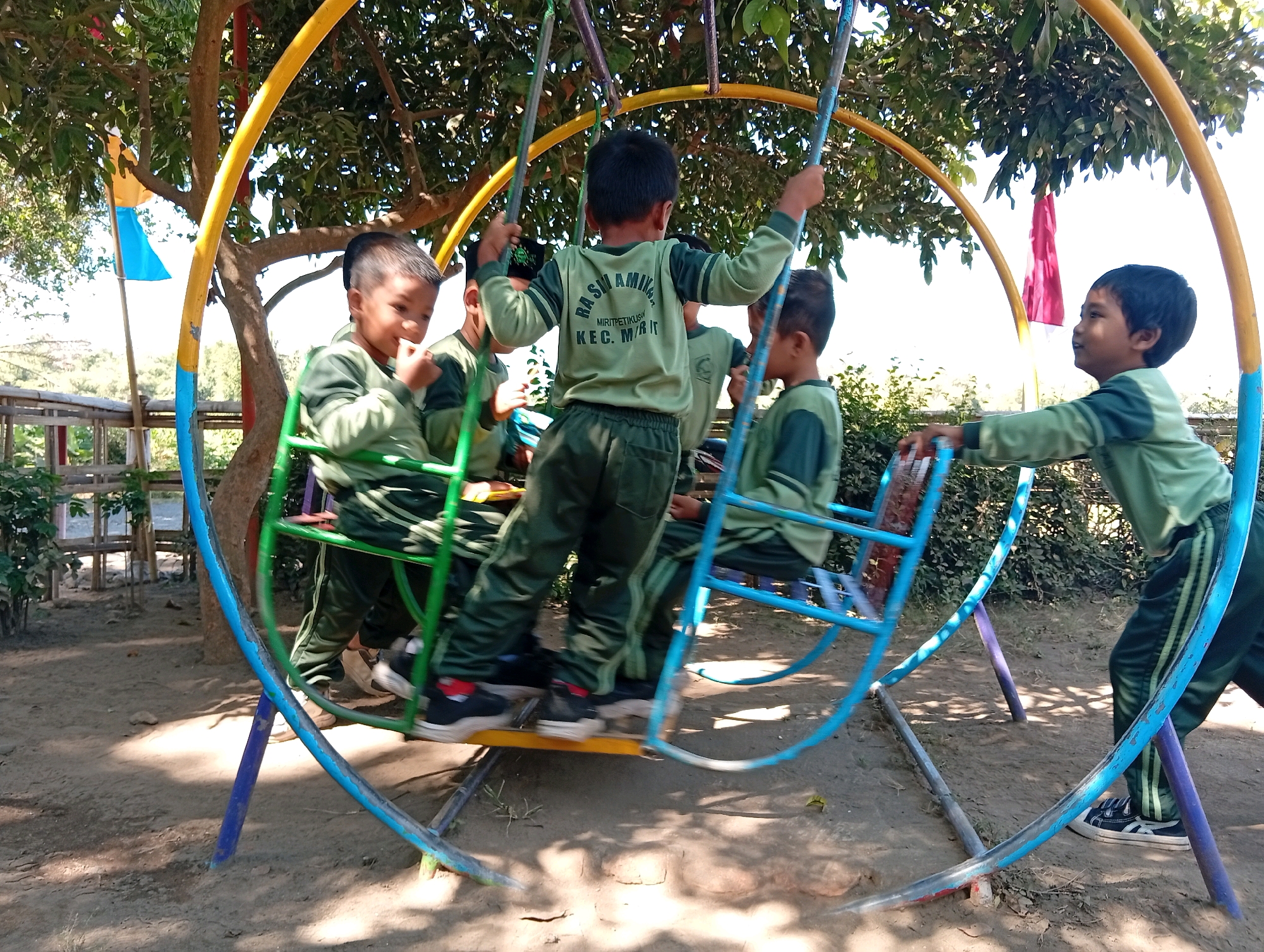 Four young boys in green uniforms with dark green stripes are playing on a multi-person swing set outdoors. The swing set has a large circular frame made of yellow and blue metal poles, with vertical purple poles supporting it. The seats of the swing are green. Two boys are on the left side of the swing, one is looking forward and smiling, while the other is looking down. On the right side, two boys are visible. One boy in the center is facing away from the camera and appears to be pushing the swing. Another boy on the far right is looking to the left. The ground is dirt, and there are trees and foliage in the background. A small blue and yellow flag is visible on a pole to the left of the swing. The overall scene suggests a schoolyard or playground environment in Miritpetikusan, Indonesia, on a sunny day.