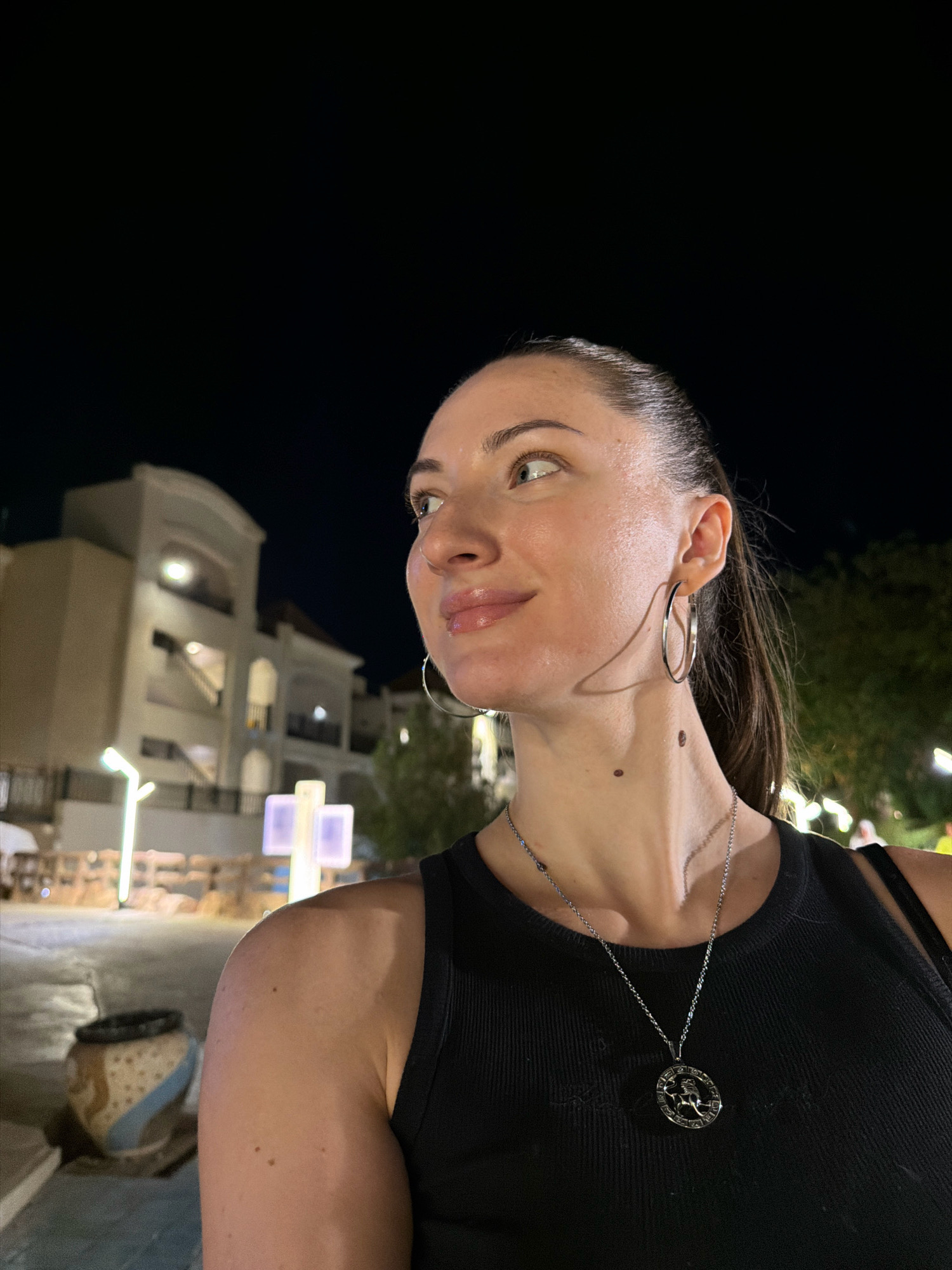 A medium shot, taken at night, shows a woman with dark hair pulled back in a ponytail. She is wearing a black tank top and silver hoop earrings. A silver pendant necklace with a circular design is visible on her neck. She has a mole on her neck and several moles on her right arm. Her skin appears dewy. She is looking to her left. In the background, a multi-story building with arched windows and balconies is illuminated. There are also lights, trees, and decorative planters visible. The scene suggests an outdoor urban or resort setting.