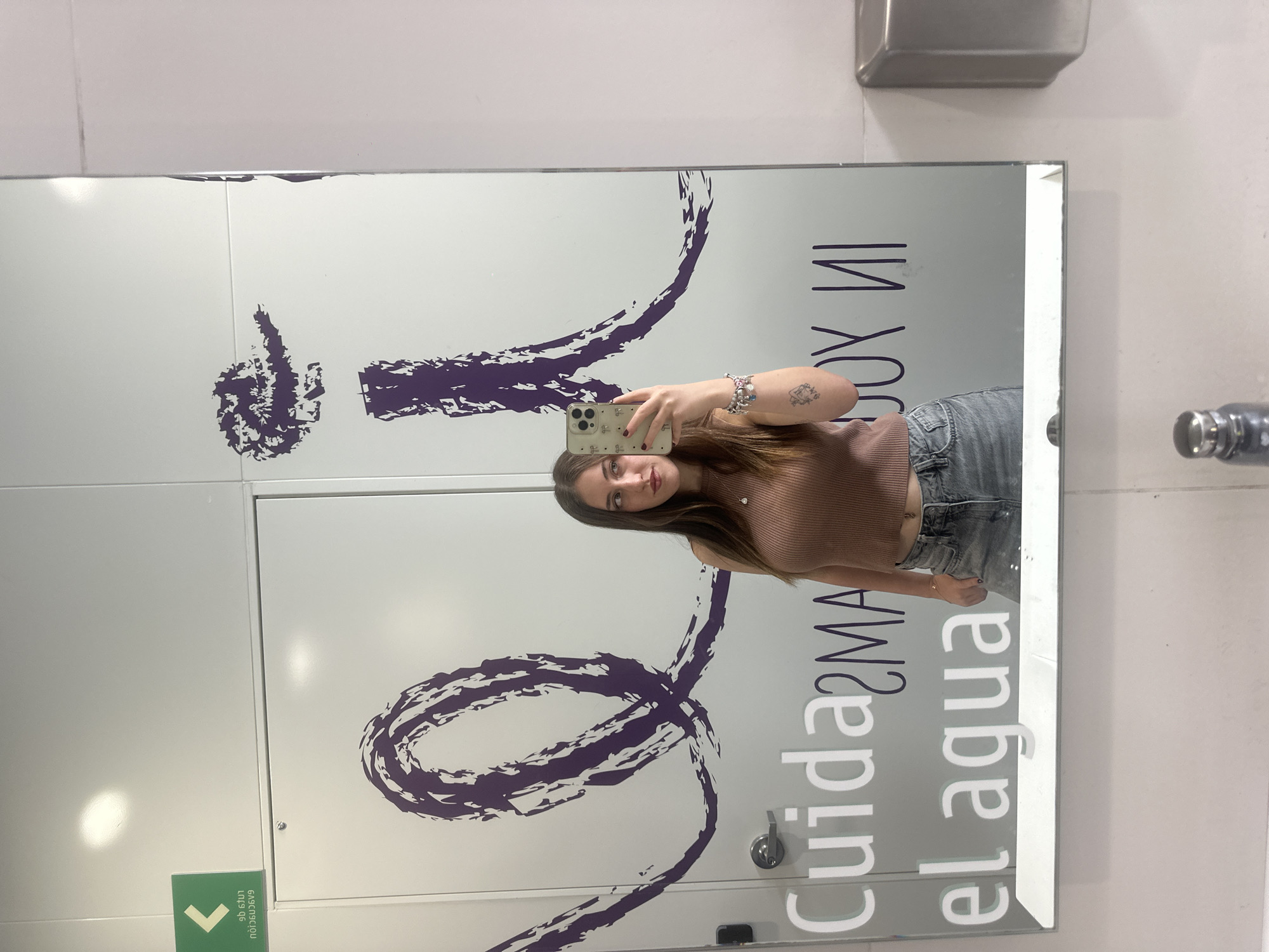 A young woman is taking a selfie in a mirror. The mirror is reflecting a restroom door. The restroom door has a purple graphic design of what appears to be water swirling, and the words "Cuida el agua" in a stylized font. There is also text above the swirling water design, but it is backwards and unreadable. A sign with a green arrow pointing left is visible at the bottom of the door. The faucet and a metallic object are seen on the white wall next to the mirror. Based on the phrase "Cuida el agua" which translates to "Take care of the water", it is likely to be in Mexico, maybe Mexico City based on the request, but this can't be verified directly in the image.