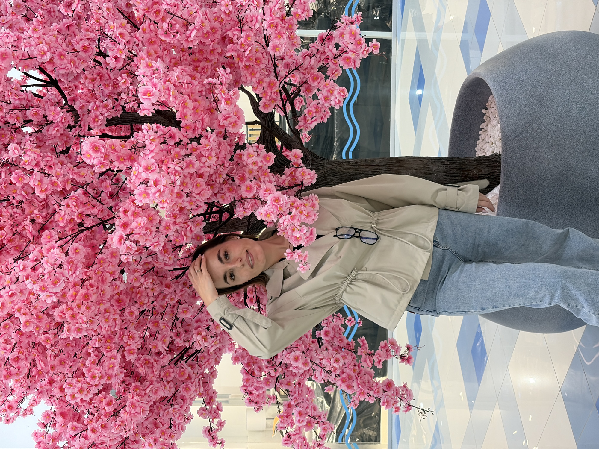 A woman poses beneath a cherry blossom tree inside a building, likely a shopping mall or commercial space in Izhevsk, Russia. She is wearing a light-colored jacket, blue jeans, and has a pair of glasses hanging from her jacket. Her left hand is near her head. The tree is in full bloom with vibrant pink flowers. The background includes a gray planter filled with white stones, a window, and a blue and white geometric tiled floor. The photo is rotated clockwise.