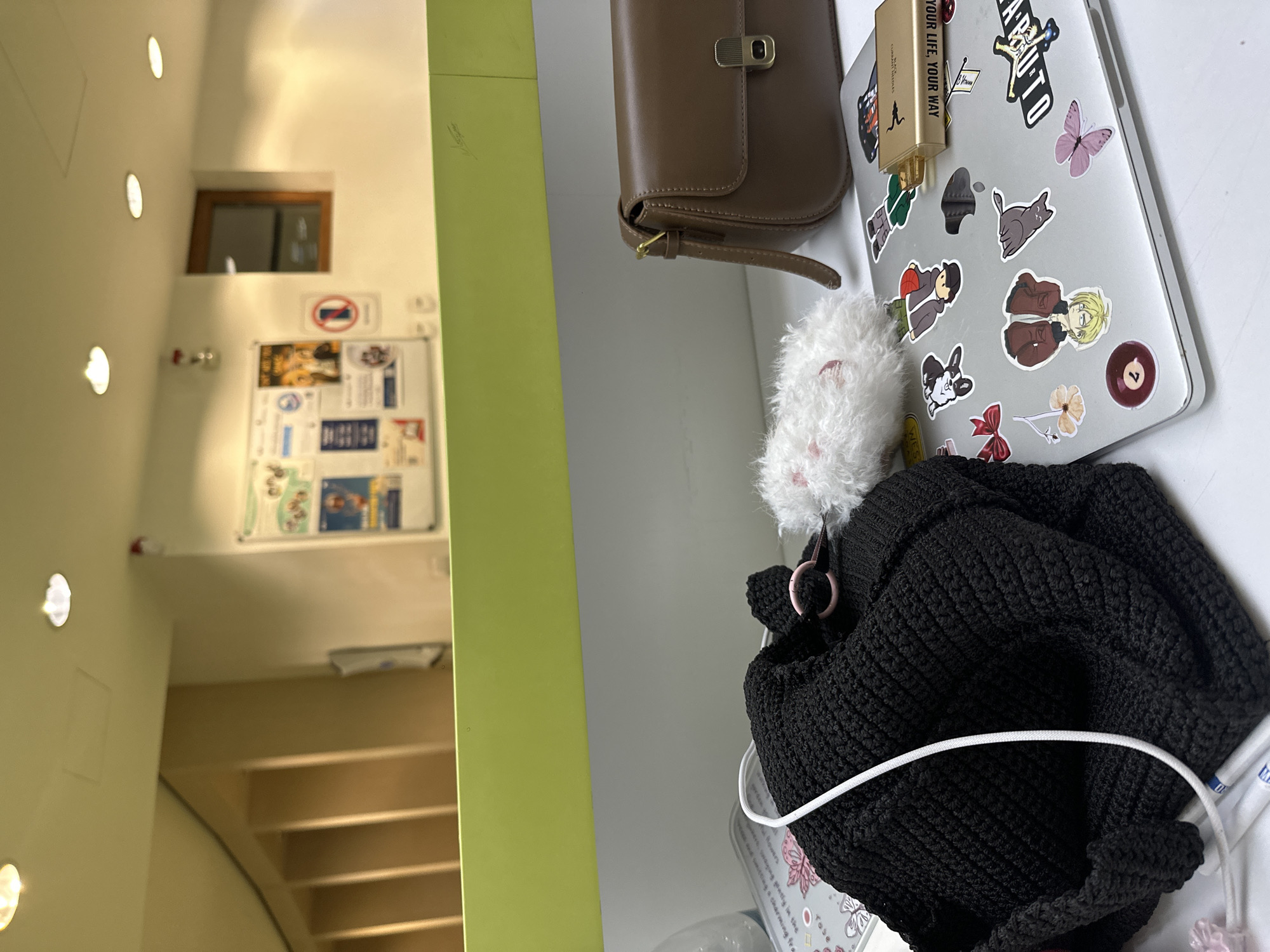 The image shows a desk with various items on it, including a brown purse, a laptop with stickers, a white fuzzy object, and a black knitted object with a white cable. The laptop has stickers of various figures and designs, including an anime character, a cat, and a butterfly. In the background, there is a hallway with lights on the wall, a bulletin board with notices, and a glimpse of stairs. The image does not provide enough information to identify the specific location in Yerevan, Armenia.