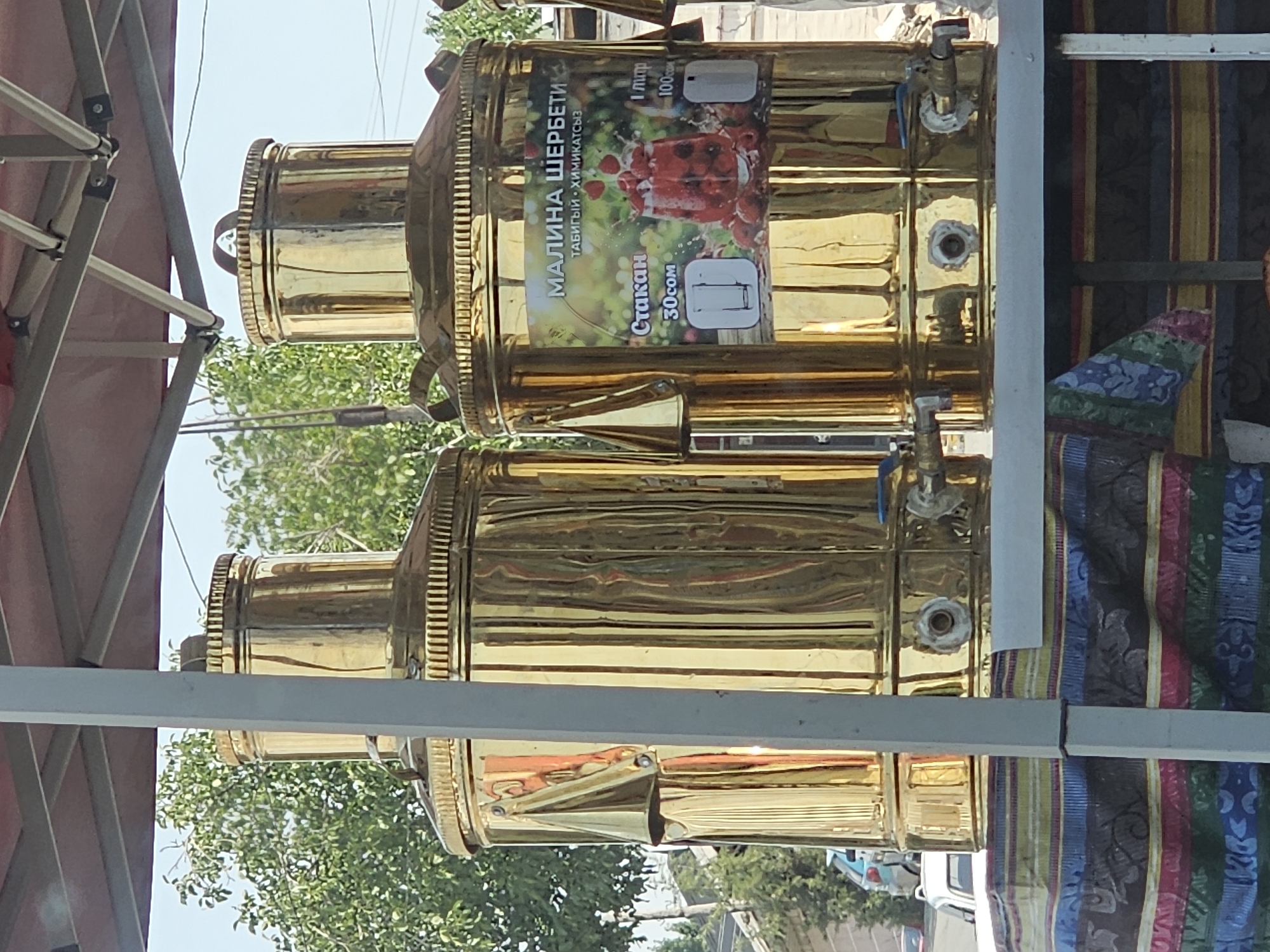 Two large, polished brass urns stand side-by-side, likely for dispensing beverages, in what appears to be an outdoor market or roadside stall. The urns are cylindrical with ribbed tops and sides, and each has a spigot near the base. One urn displays a colorful label featuring raspberries and a glass of raspberry drink, with text in Cyrillic, indicating it dispenses "МАЛИНА ШЕРБЕТИ" (Raspberry Sherbet). The price for a glass is listed as 30som, and for 1 liter as 100som. The urns are situated on a table covered with a vibrant, patterned textile. In the background, trees with green foliage are visible against a clear sky, suggesting a daytime setting. The presence of vehicles and buildings in the blurred background hints at an urban or semi-urban environment. The city of Özgön in Kyrgyzstan is a plausible location for such a scene, given the prevalence of outdoor markets and the style of the beverages and their presentation.