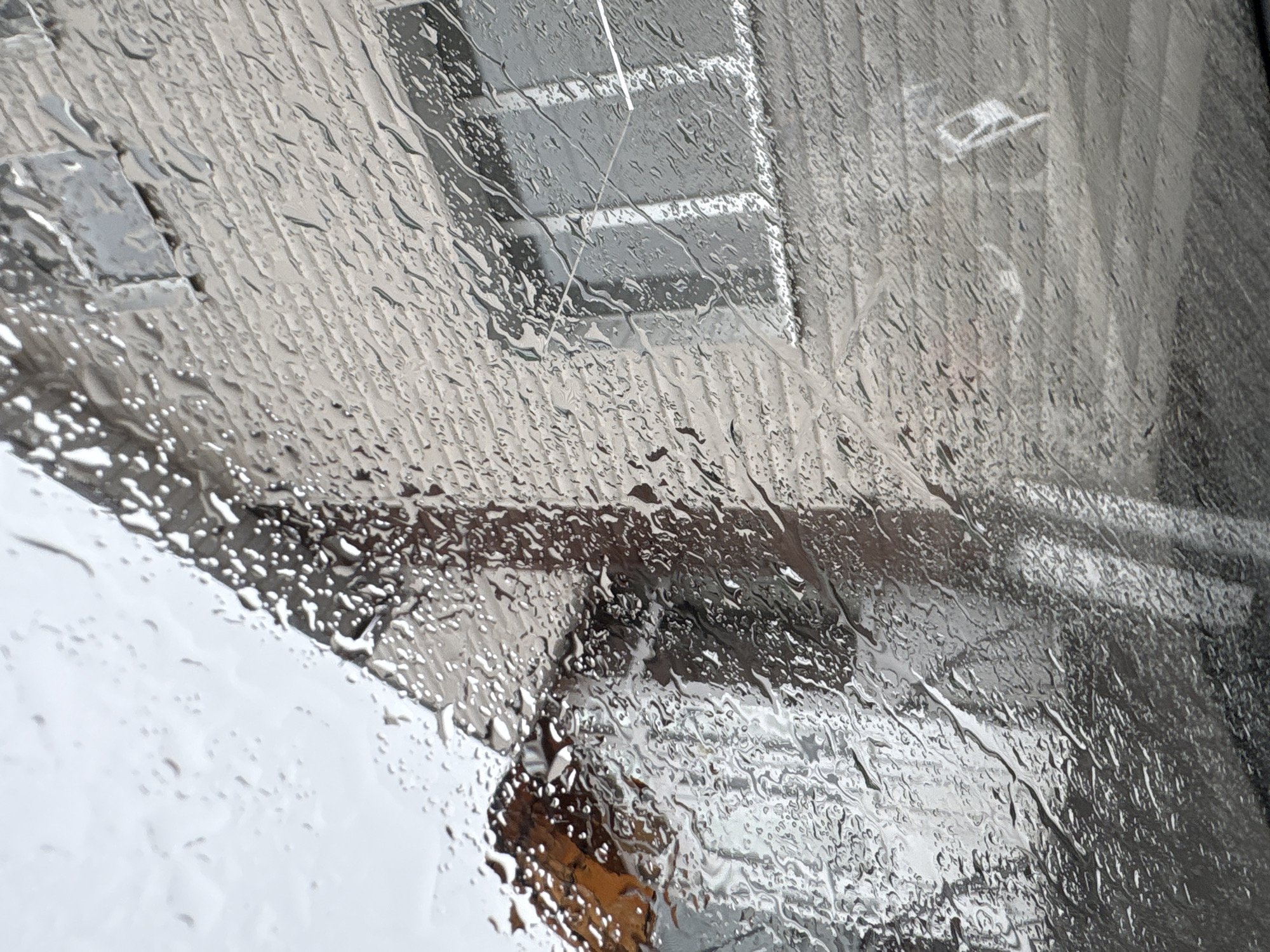 The image captures a close-up, distorted view through a window covered in numerous raindrops. The foreground is dominated by water droplets and streaks of varying sizes, which refract and blur the external scene.
Through the rain-streaked glass, a greyish, likely urban or suburban environment is visible. The primary background element appears to be a building with vertical textured details, possibly siding or paneling. In the upper-middle section, a rectangular window frame or a similar architectural detail with horizontal divisions can be vaguely discerned through the water. Further to the right, indistinct shapes suggest more of the structure or adjacent buildings.
The bottom-left portion of the image shows a bright, blurry white area, indicating the presence of snow on the ground, which, along with the rain, points to cold and wet weather conditions. A dark, indistinct brownish-orange shape is visible in the lower-middle, possibly an object or reflection, but its identity is obscured by the water.
The scene conveys a sense of introspection or coziness indoors while observing inclement weather outside during daytime. No specific activity is depicted, and no people or visible text are present. The overall atmosphere is muted and somewhat melancholic due to the heavy rain and grey tones, against the provided location context of Прикамский, Russia.