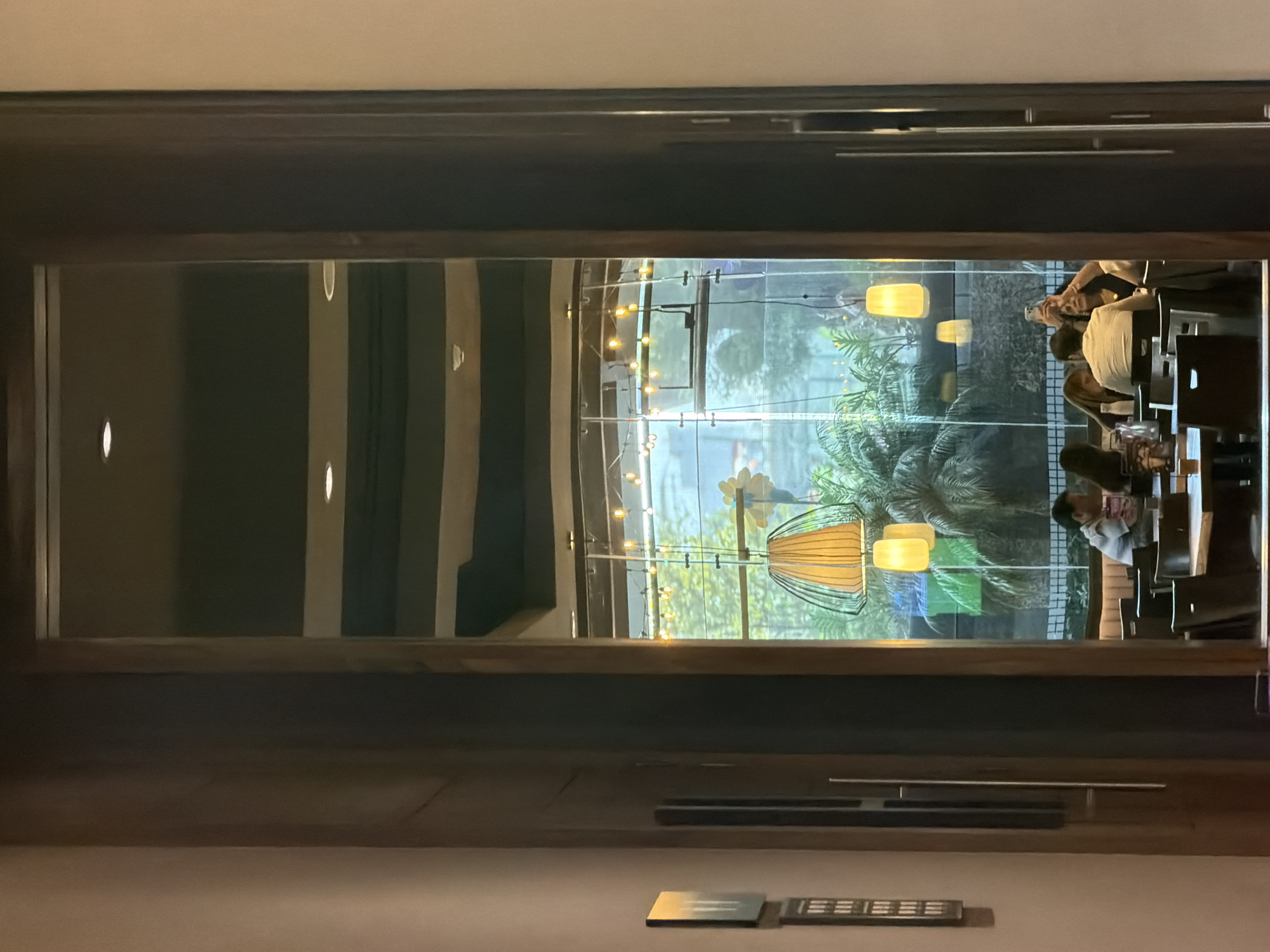 The image, oriented vertically, captures an interior view looking out through a large, framed window or glass partition. The setting appears to be a restaurant or cafe in Mexico City, Mexico.
In the foreground, on the interior side, a dark wooden frame encloses the clear glass. Below the window, a dark surface is visible, along with a horizontal silver bar. Further down, near the bottom of the frame, there's a small, flat metallic object and what appears to be a remote control with a grid of small buttons or indicators. No readable text is clearly discernible on these items.
Beyond the glass, an exterior scene unfolds, characterized by lush green foliage, likely palm trees or similar tropical plants. The ambiance outside is brighter than the interior, suggesting natural light. Several decorative light fixtures are visible: some are yellow or amber cylindrical pendant lights, while another is a larger, cage-like lantern with a warm glow. A string of small, round fairy lights stretches horizontally across the view, adding to the decorative illumination.
Several people are seated at tables beyond the glass on the right side of the image. One individual in a light-colored shirt is visible, looking downwards. Another person's arm is raised, holding a smartphone, possibly capturing a photo or video of the surroundings. The general activity appears to be dining or socializing in a relaxed setting. The presence of artificial lighting suggests it might be evening or night, or an interior designed for a warm, intimate atmosphere. Reflections of interior lights and possibly other structural elements are visible on the glass.