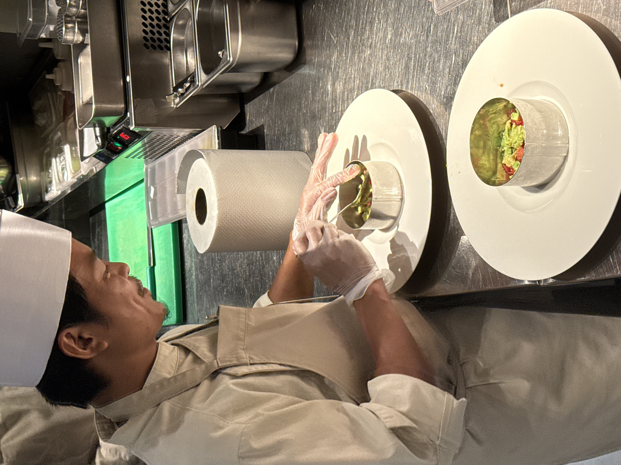 A male chef, wearing a white chef's hat and a light-colored chef's uniform with an apron, is meticulously preparing food in a commercial kitchen. He is standing at a stainless steel counter and is in the process of plating a dish. The chef is wearing clear plastic gloves and is using a spoon to arrange ingredients within a circular metal ring on a white plate. The dish appears to contain finely diced vegetables, possibly avocado and tomatoes.
In the background, there are various kitchen elements: a bright green cutting board, stainless steel containers, a roll of paper towels, and what appears to be a piece of kitchen equipment with a digital display. The overall environment suggests a professional culinary setting. The image captures a close-up view of the chef's hands and the food preparation process. The lighting suggests an indoor environment, likely a restaurant kitchen. Without specific visual cues indicating Dubai, United Arab Emirates, this location cannot be confirmed.