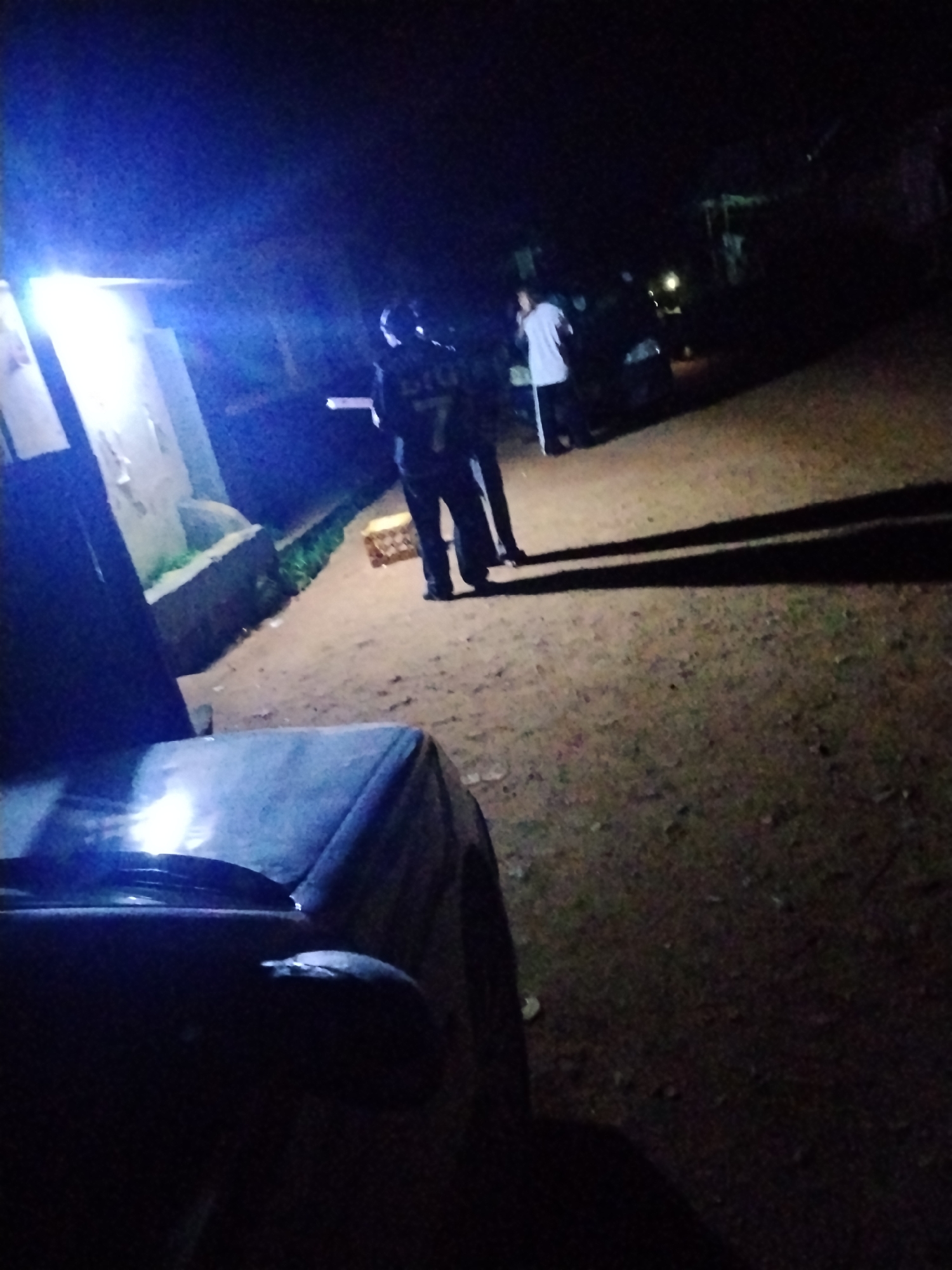 In Wumat, Nigeria, at night, a group of people are visible near a building. One individual, wearing a dark uniform with the number "7" and lettering "BIG" on the back, stands facing away from the camera. Another person in a white shirt stands near a dark-colored car. The ground appears to be dirt, and there's a light source to the left, casting a bright blue glow. A portion of a dark-colored vehicle is in the foreground, partially obscuring the view. The scene suggests an outdoor gathering or activity taking place in a dimly lit, possibly rural or semi-urban setting.