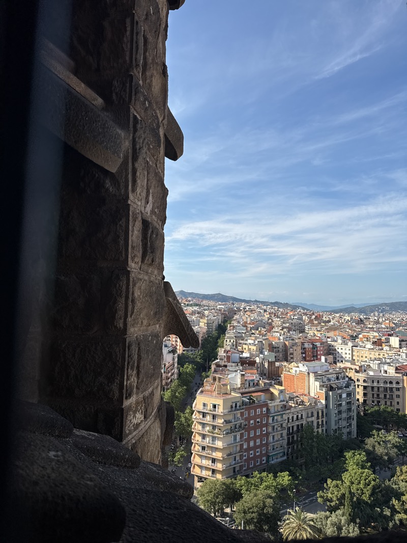 This image captures an expansive, elevated view of a city sprawling under a bright blue sky. A large, dark, weathered stone architectural element dominates the left and bottom foreground, acting as a partial frame for the urban panorama. This stone structure, characterized by its rough, rectangular blocks and distinct carved protrusions, suggests it is part of a significant, possibly historic, building or tower.
Beyond the foreground, the city unfolds with numerous multi-story buildings, many featuring balconies and varied architectural styles. Streets lined with green trees create clear avenues, and patches of foliage, including palm trees, are visible throughout the urban fabric. In the distance, low-lying hills or mountains define the horizon.
The scene is bathed in clear, bright sunlight, indicating a late morning or early afternoon time of day. The sky is largely clear, with only a few wispy white clouds. No specific human activity or people are distinctly visible. The characteristic urban layout, architecture, and distant hills strongly suggest a Mediterranean city, possibly Barcelona, Spain, viewed from a high vantage point, such as a famous landmark structure. There is no visible text in the image.