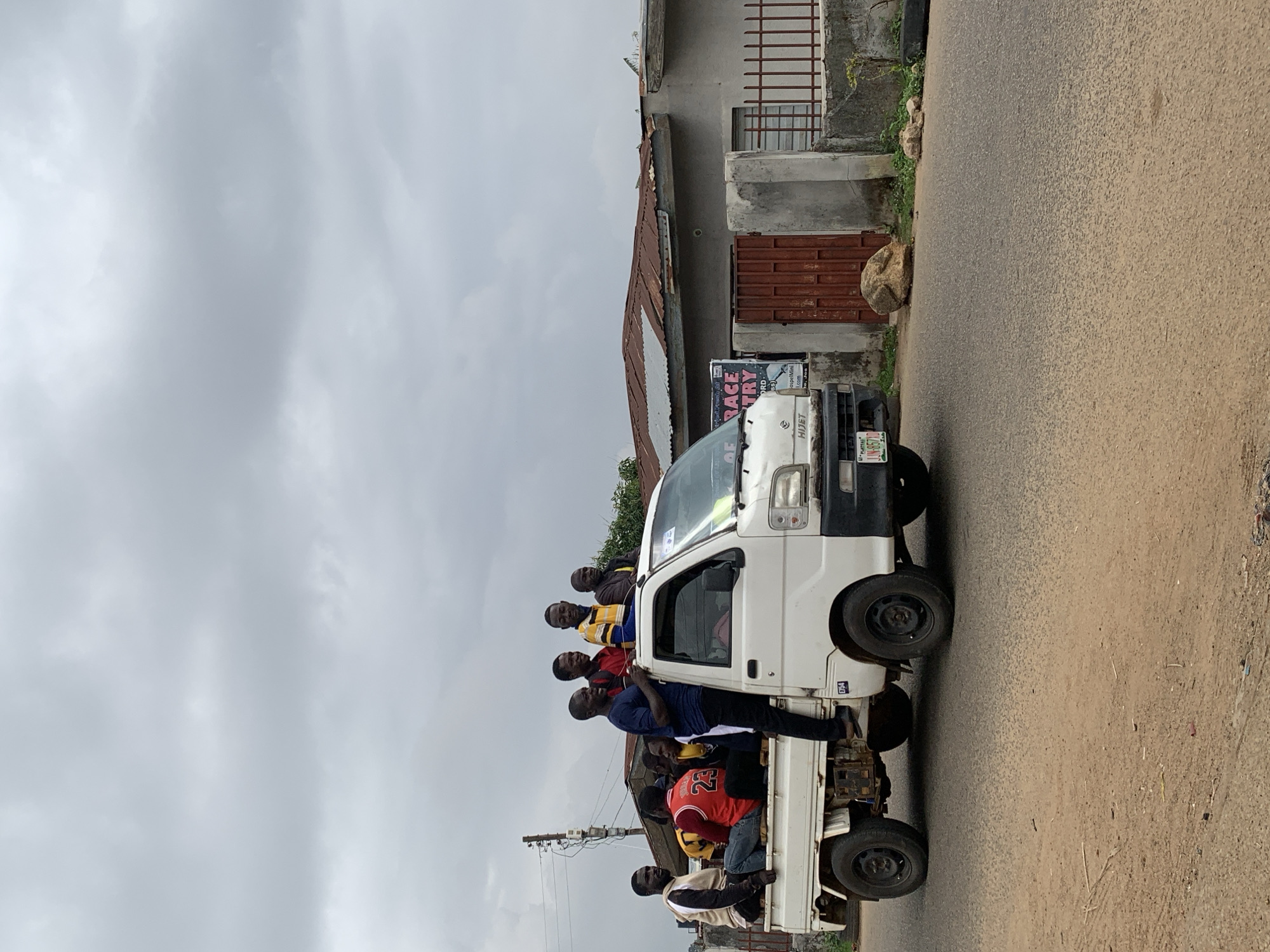 The image depicts a white Daihatsu Hijet mini truck overloaded with at least eight individuals. Some passengers are inside the cab, while others are riding on the bed of the truck. The truck is traveling on an asphalt road, with an overcast sky overhead. A building with a weathered metal roof and a rusty metal gate is visible in the background, along with a sign that reads "of Grace Race Ministry". The license plate of the vehicle is visible, which gives the location as Jos, Nigeria. The overall scene suggests transportation of people in a manner that may exceed safe carrying capacity.