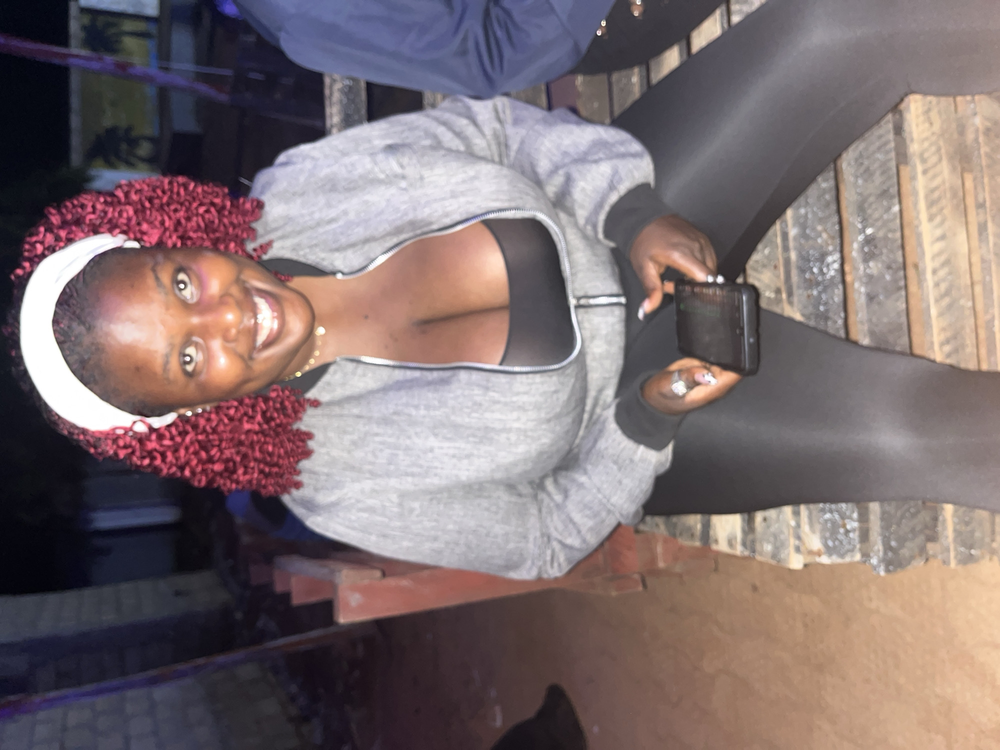 A woman with dark skin, bright red curly hair, and a white headband smiles at the camera while sitting on a wooden bench. She is wearing a gray zip-up jacket over a black top that reveals her cleavage, and black leggings. She holds a smartphone in her right hand. The setting appears to be outdoors at night, possibly in Durumi, Nigeria, with a brick wall visible behind her and part of another person to her right. The ground is a mix of dirt and what looks like wooden pallets.