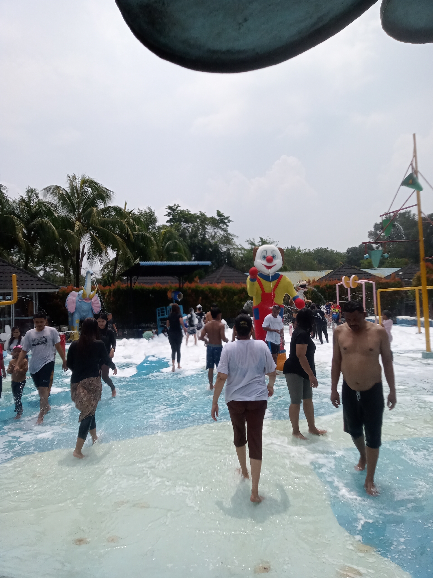 The image depicts a busy water park scene in Baru, Indonesia, with many people enjoying the foam and water. In the center, a large, inflatable clown character stands prominently. The water is a bright blue and white, indicating a shallow pool or splash area filled with foam. Palm trees and tropical foliage are visible in the background, suggesting a warm climate. There are also some structures resembling cabanas and water park attractions. People of various ages are dressed in swimwear and casual clothing, with some wading through the water and others walking around. The atmosphere appears lively and recreational.