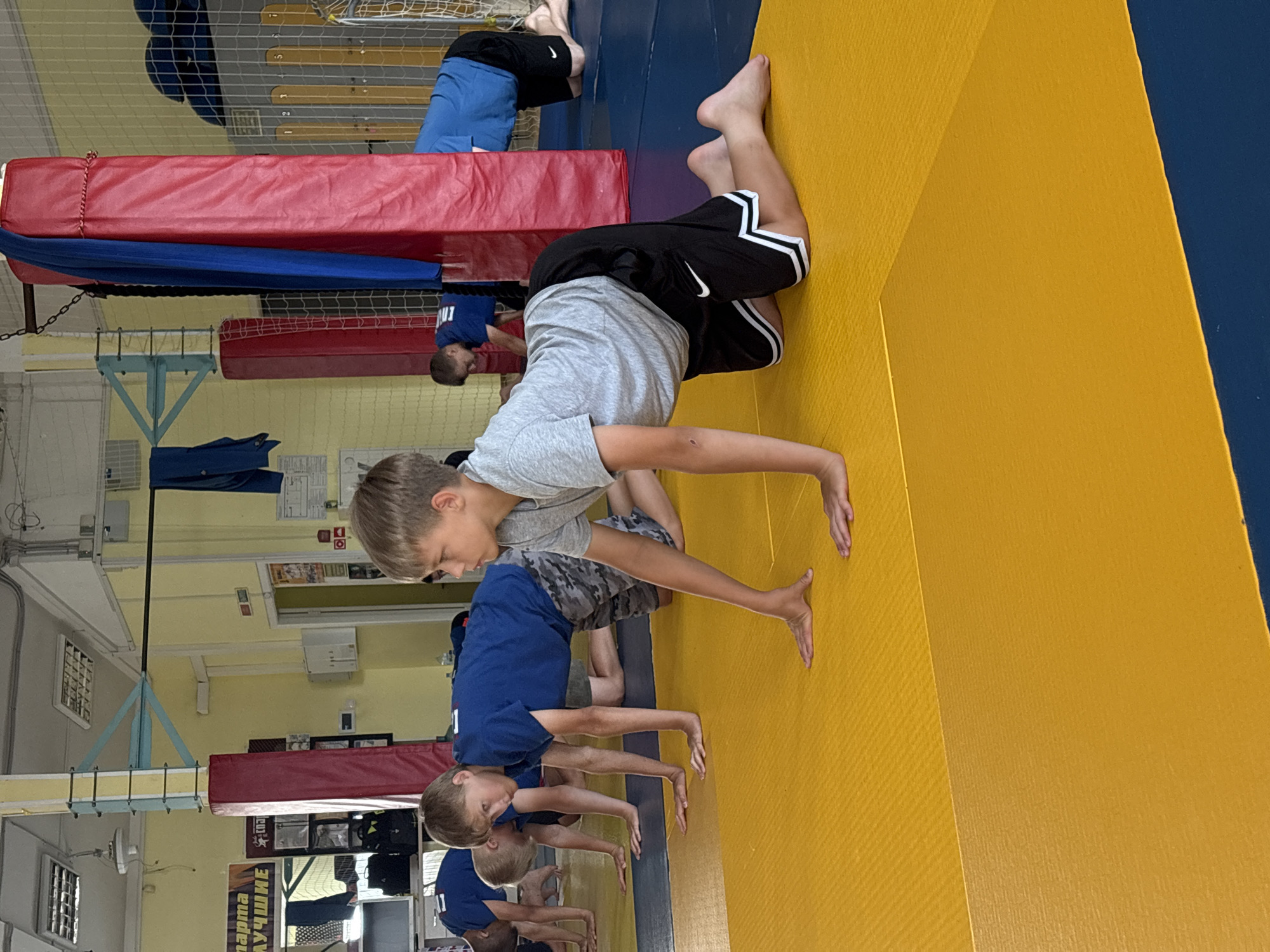 The image shows a group of young boys in athletic attire performing an exercise on yellow and blue mats in what appears to be a gymnasium or sports hall. The boys are on their hands and knees, with their arms extended. The room has a yellow-painted wall with blue and red padded pillars, and netting in the background. Posters and sports equipment are visible in the background. The overall scene suggests a sports training or practice session. The location is identified as Chaikovsky, Russia, likely a wrestling or martial arts training facility given the mats and padded pillars.