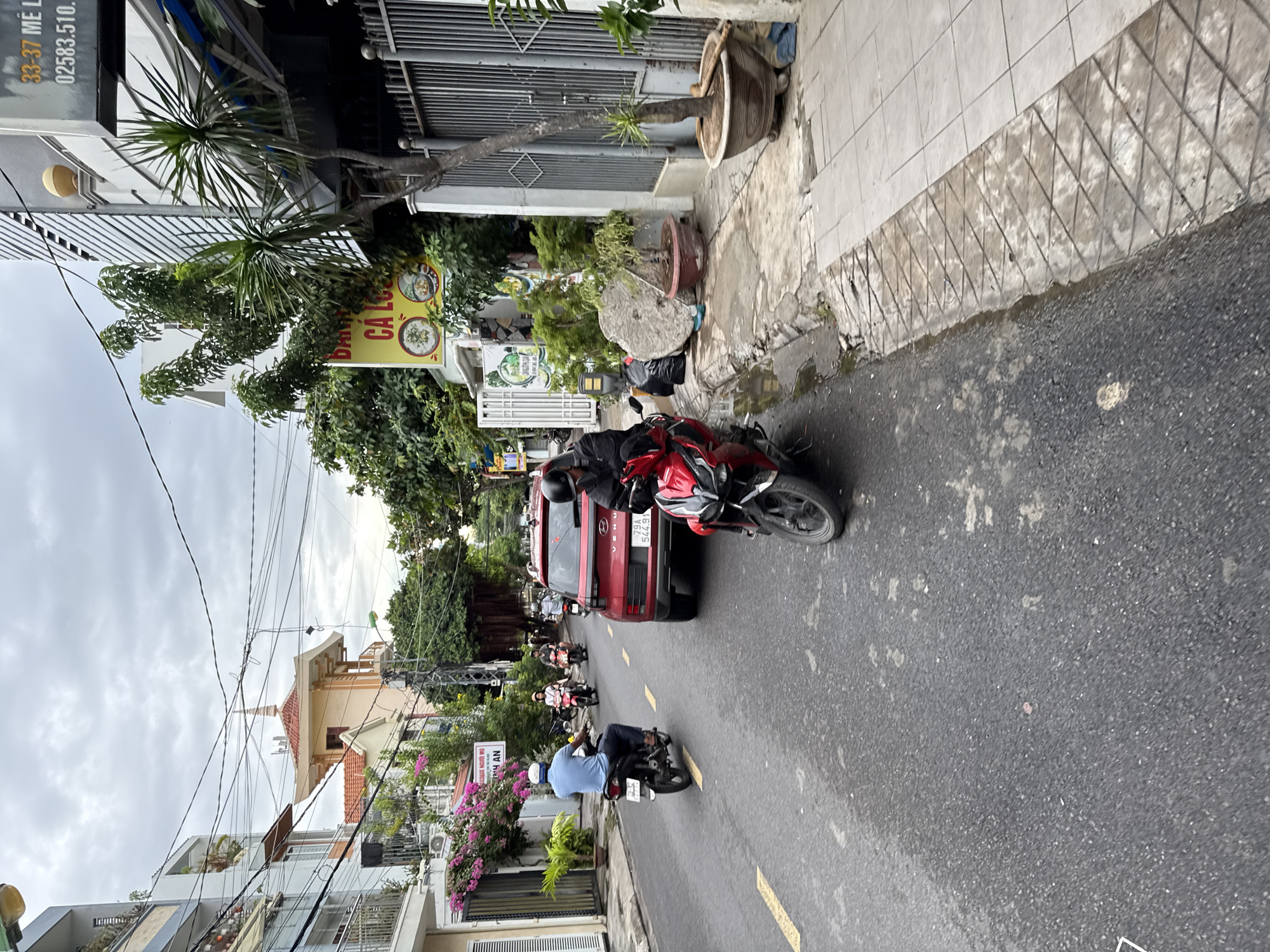 This image captures a street scene in Nha Trang, Vietnam, likely in a commercial or residential area, given the buildings and road infrastructure. The sky is overcast, suggesting a cloudy day.
In the foreground, a red scooter is parked on the right side of the road, with its front wheel turned slightly. A person wearing a black jacket and helmet is seated on this scooter, looking towards the left. Further down the street, a red Hyundai SUV with license plate "79A 544.91" is stopped, facing away from the camera.
To the left of the road, a man wearing a blue shirt and dark pants is riding a black scooter away from the camera. He is wearing a white helmet. Several other individuals on scooters are visible further down the street in the background, moving in the same direction.
The buildings lining the street are a mix of styles. On the left, modern multi-story buildings with balconies and windows are visible, some adorned with flowering plants. On the right, a building with a metal fence and a tree in a pot is present. A yellow sign with Vietnamese text and images of food is attached to a building on the right. Power lines crisscross overhead.
The road surface appears to be asphalt, with some dampness and scattered debris. The sidewalk on the right has a tiled pattern and a raised curb. The overall impression is of a busy urban street with active traffic.