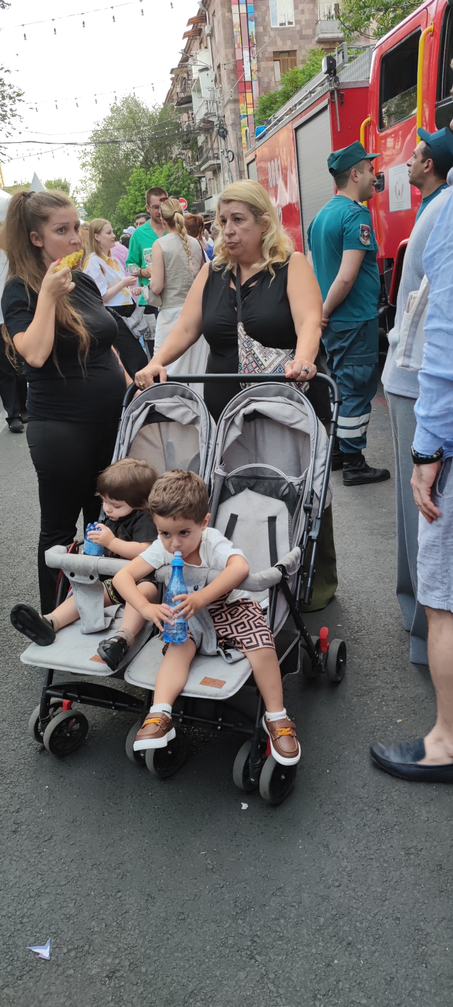 This detailed image captures an outdoor street scene in Yerevan, Armenia, during what appears to be a public event or festival.
In the immediate foreground, two young boys are seated side-by-side in a light grey and black double stroller. The boy on the left, with dark hair, wears a black t-shirt and dark patterned shorts with black sandals, looking downwards while holding a small blue water bottle with a white cap. The boy on the right, with lighter brown hair, wears a white t-shirt and brown shorts with a black geometric (Greek key style) pattern, white socks, and brown and orange velcro shoes. He is actively drinking from a larger blue water bottle with a white sippy top, looking directly towards the viewer. The asphalt pavement beneath the stroller shows minor debris, including a small piece of folded white paper.
Behind the stroller, two women are partially visible, interacting with it. The woman on the right, pushing the stroller, has shoulder-length blonde hair and wears a black sleeveless top, a ring on her right hand, and a colorful patterned cross-body bag. She has a neutral expression. To her left, another woman with long brown hair, wearing a black fitted t-shirt and black pants, appears to be pregnant, with a visible belly. She is actively eating a yellow food item, possibly grilled corn or a kebab, holding it to her mouth with her right hand while her left hand rests on the stroller's handle.
In the midground, a diverse crowd of people can be seen, some holding glasses, suggesting a social gathering. String lights are visible strung overhead across the street.
The background features a large red fire truck with "0001" visible on its side, parked parallel to the street. Two men in teal/blue-green uniforms and matching caps, presumably firefighters or emergency personnel, are standing near the truck, engaged in conversation. Behind them, multi-story stone buildings with traditional Armenian architectural elements, including ornate balconies and windows, line the street. One building has a vertical decorative mosaic or tile pattern on its facade. The sky above is bright and appears overcast.