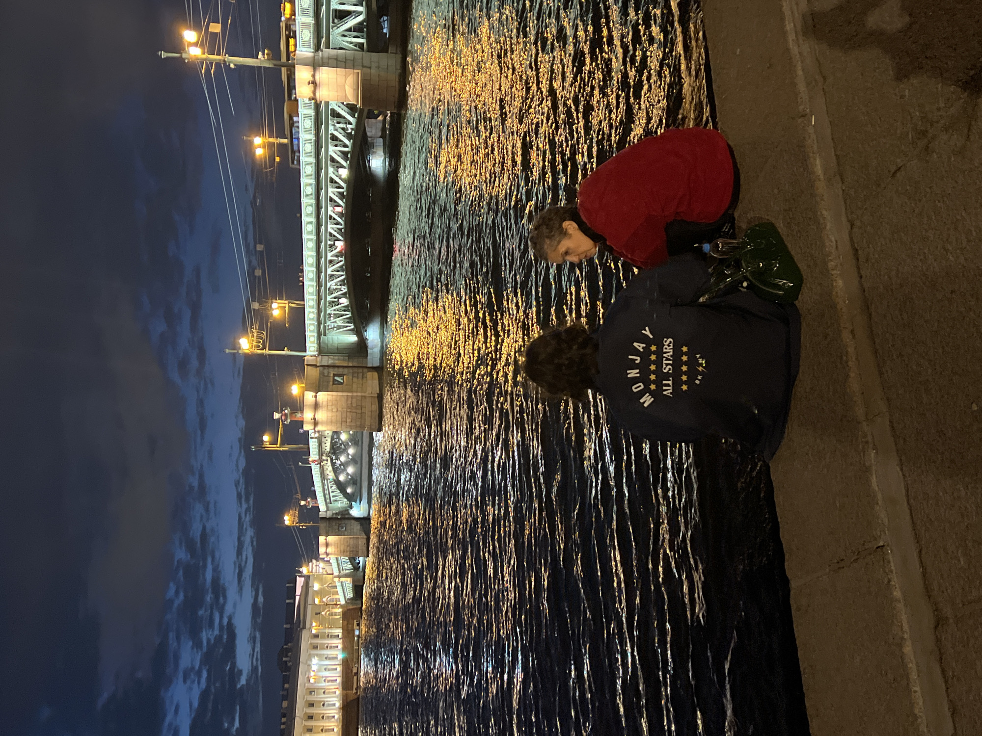 Two individuals are seated on a concrete embankment facing a body of water at twilight. The person on the left, viewed from the back, wears a dark blue jacket with white text and stars on the back, reading "MONJAY ALL STARS". A green handbag and a clear plastic bottle are beside them. The person on the right, facing left, is wearing a red jacket. The water reflects the lights of a bridge and buildings in the background. The bridge, illuminated by numerous lights, spans the water, with architectural details visible. A large building with many windows and lights is situated to the left of the bridge. The sky is dark blue with scattered clouds, and the scene appears to be Saint Petersburg, Russia, likely along the Neva River, given the presence of a bridge and substantial waterfront architecture common to the city.