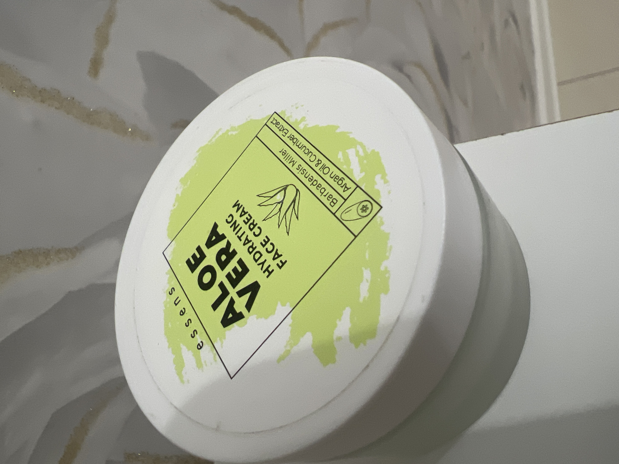 A white cylindrical tub of "essens ALOE VERA HYDRATING FACE CREAM" is shown. The tub has a light green label with black text and an illustration of an aloe vera plant. Below the aloe vera illustration, there is text that reads "Barbadensis Miller Argan Oil & Cucumber Extract." The tub is positioned on a surface, likely a countertop or shelf, with a textured, light-colored wall in the background. No people are visible. The image is taken from a slightly elevated angle, focusing on the top of the tub. There is no information about the city of Mannheim, Germany.