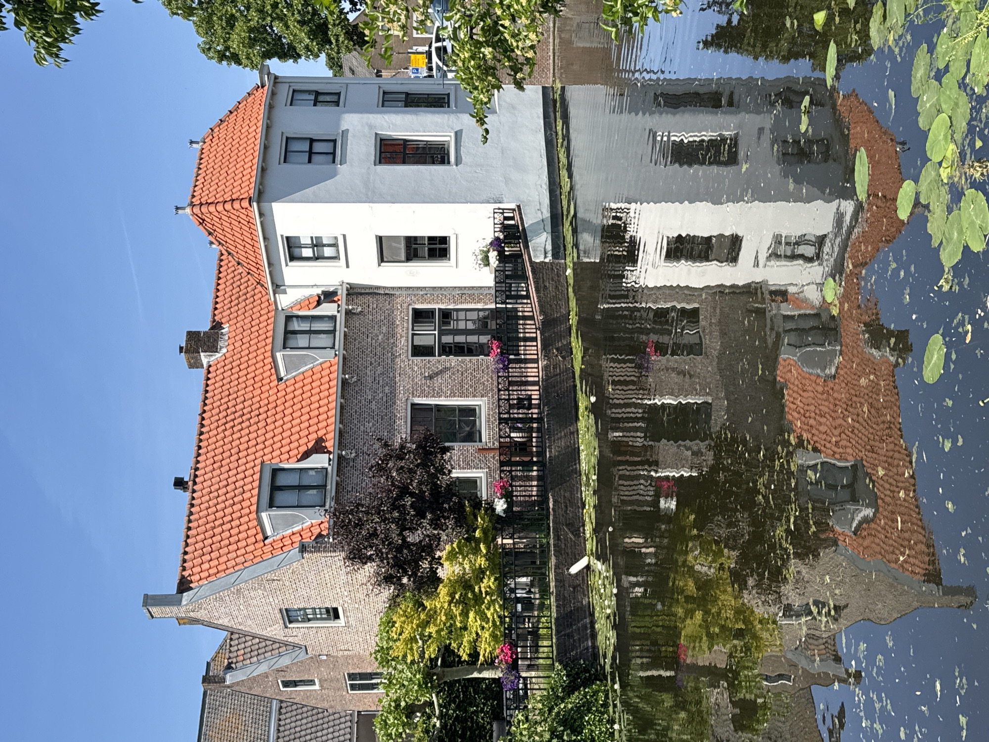 The image shows a view of buildings in Katwijk, Netherlands, reflected in the water. The architecture features a mix of brick and white facades, some with red-tiled roofs. There are trees and other vegetation along the waterfront, contributing to the scene. The water's surface reflects the buildings and vegetation, creating a distorted, mirrored image. The sky is visible in the upper left corner, appearing blue.