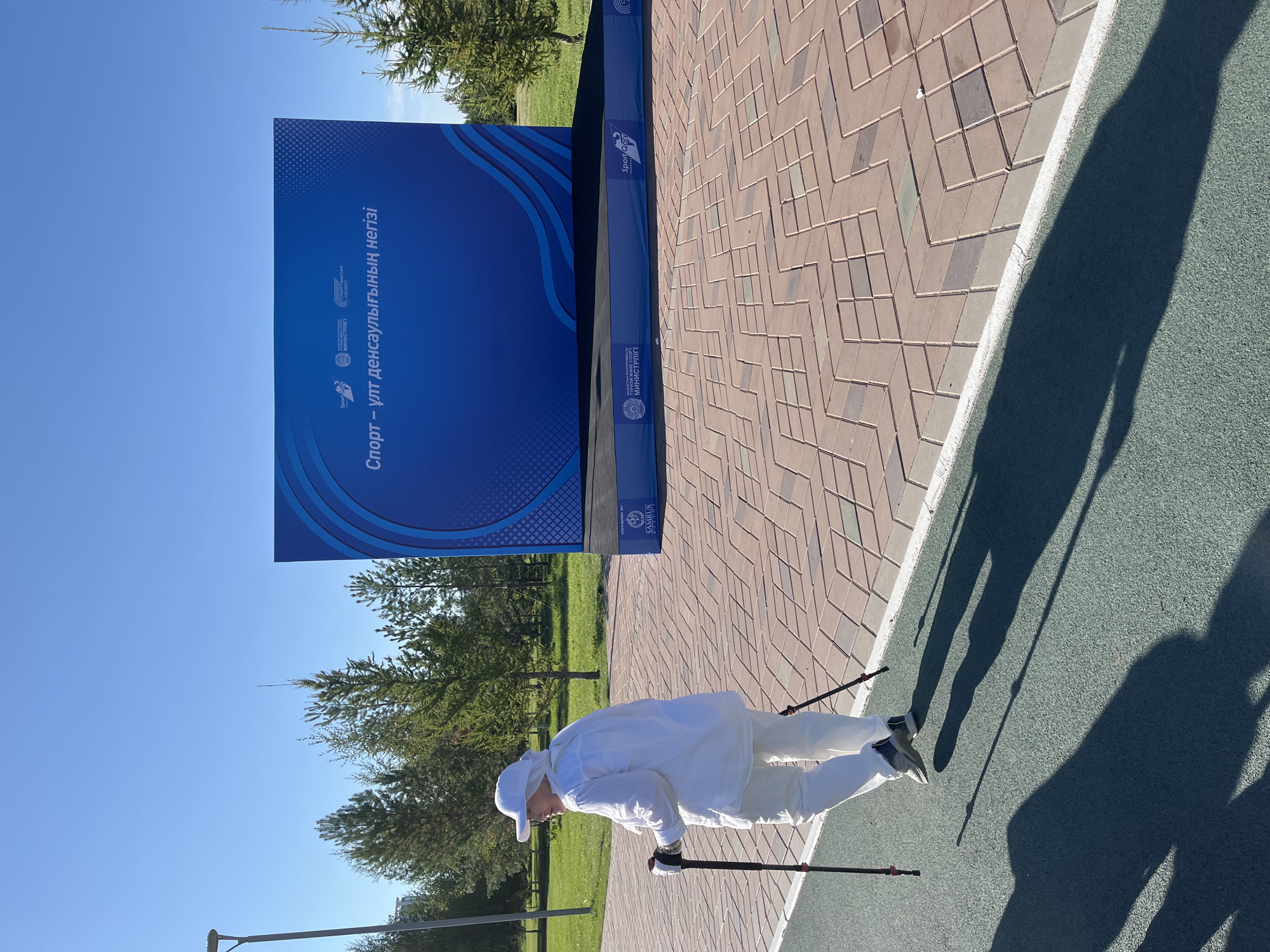 An elderly woman, dressed in white attire including a hat, is Nordic walking on a green track in a park. She is using two poles with red accents. The track is bordered by a patterned brick pathway. In the background, a large blue banner displays text in Cyrillic script, which appears to be related to sports and health. The banner is supported by a dark platform with logos, including "SPORT OTYRY" and "SAMRUK KAZYNA". Several trees are visible behind the banner, and the sky is clear and blue. The overall setting suggests an outdoor event or recreational area in Astana, Kazakhstan, likely during daytime with strong sunlight casting shadows.
