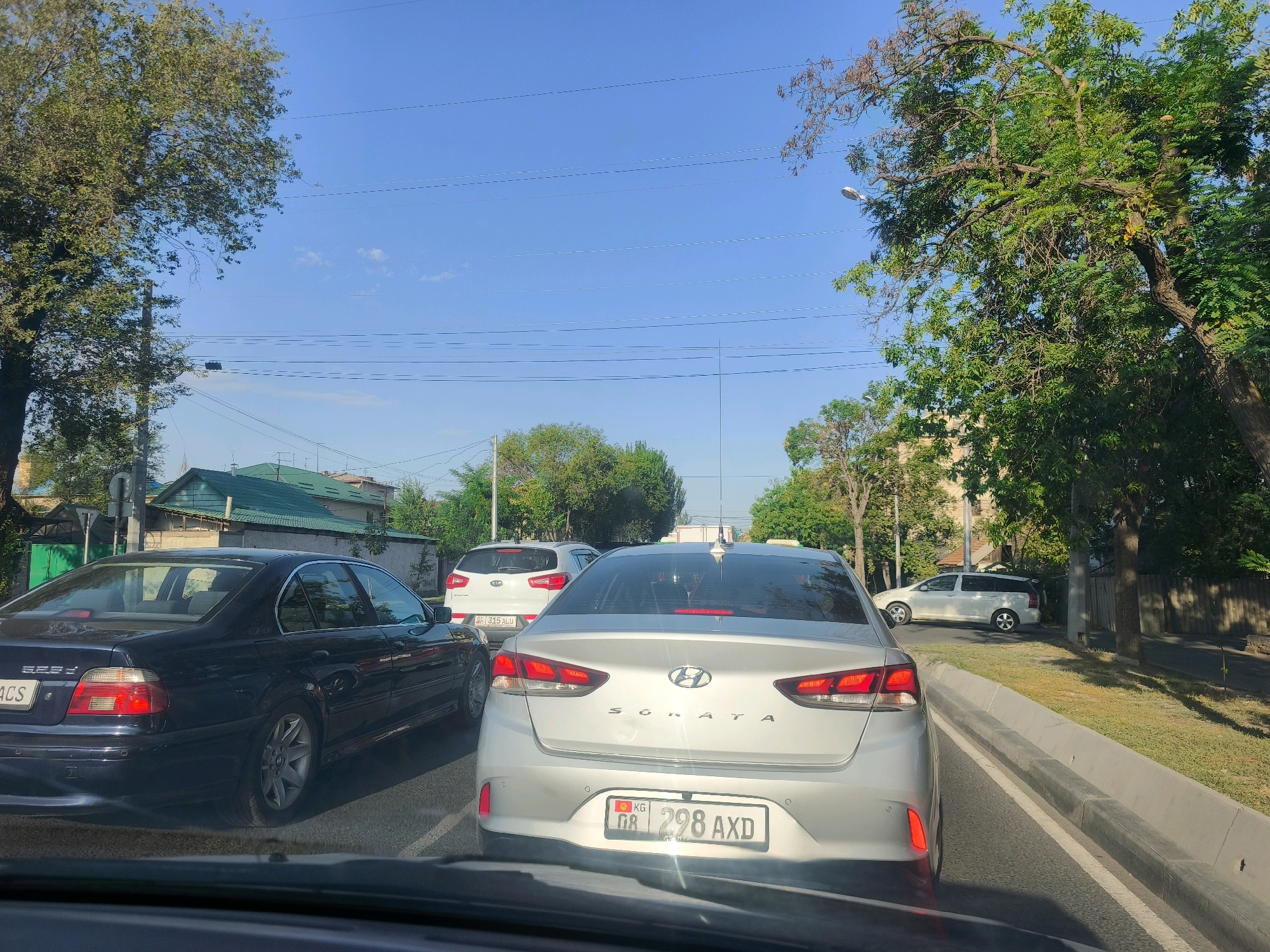 The image shows a street scene with several cars in traffic. In the foreground, the dashboard and windshield of a vehicle are visible, suggesting the photo was taken from inside a car. Ahead, a silver Hyundai Sonata is in the lane, its rear end facing the camera. To the left of the Sonata, a white Kia Sportage is also in traffic. Further left and slightly behind is a dark BMW sedan. On the right side of the street, a white minivan is parked. The background shows trees, buildings with green roofs, and a bright blue sky with power lines. The environment appears to be a sunny day in a city.