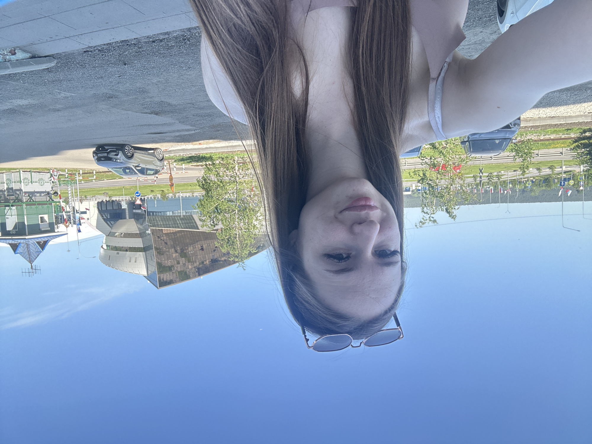 The image is rotated 90 degrees clockwise. It shows a young woman taking a selfie, with her face and upper torso in the foreground, captured from a low angle. Her head is oriented towards the bottom of the frame, and her long, straight brown hair cascades towards the left and right sides. Her eyes are open, looking slightly upwards towards the viewer, and she displays a subtle smile. Sunglasses rest on her chin, with the lenses facing away from her face. She has fair skin and is wearing a light-colored top with a delicate strap visible on her right shoulder.
In the background, along what appears to be the top of the rotated image, an outdoor urban or suburban scene unfolds. This includes a paved road and a sidewalk, with a silver/grey SUV or crossover parked on the side. The vehicle's license plate shows partially legible characters, possibly "A01" or "H01". Further behind are several buildings, notably one with a distinctive blue, peaked roof and another modern structure constructed with glass and dark panels. Green trees, bushes, and grass are scattered throughout the landscape. Traffic signs, such as a blue circular sign with a white arrow, and small, unidentifiable flags are also present. The sky, which occupies the large lower portion of the rotated image, is a bright, clear blue, indicating sunny daytime weather. No other clear, readable text is visible.