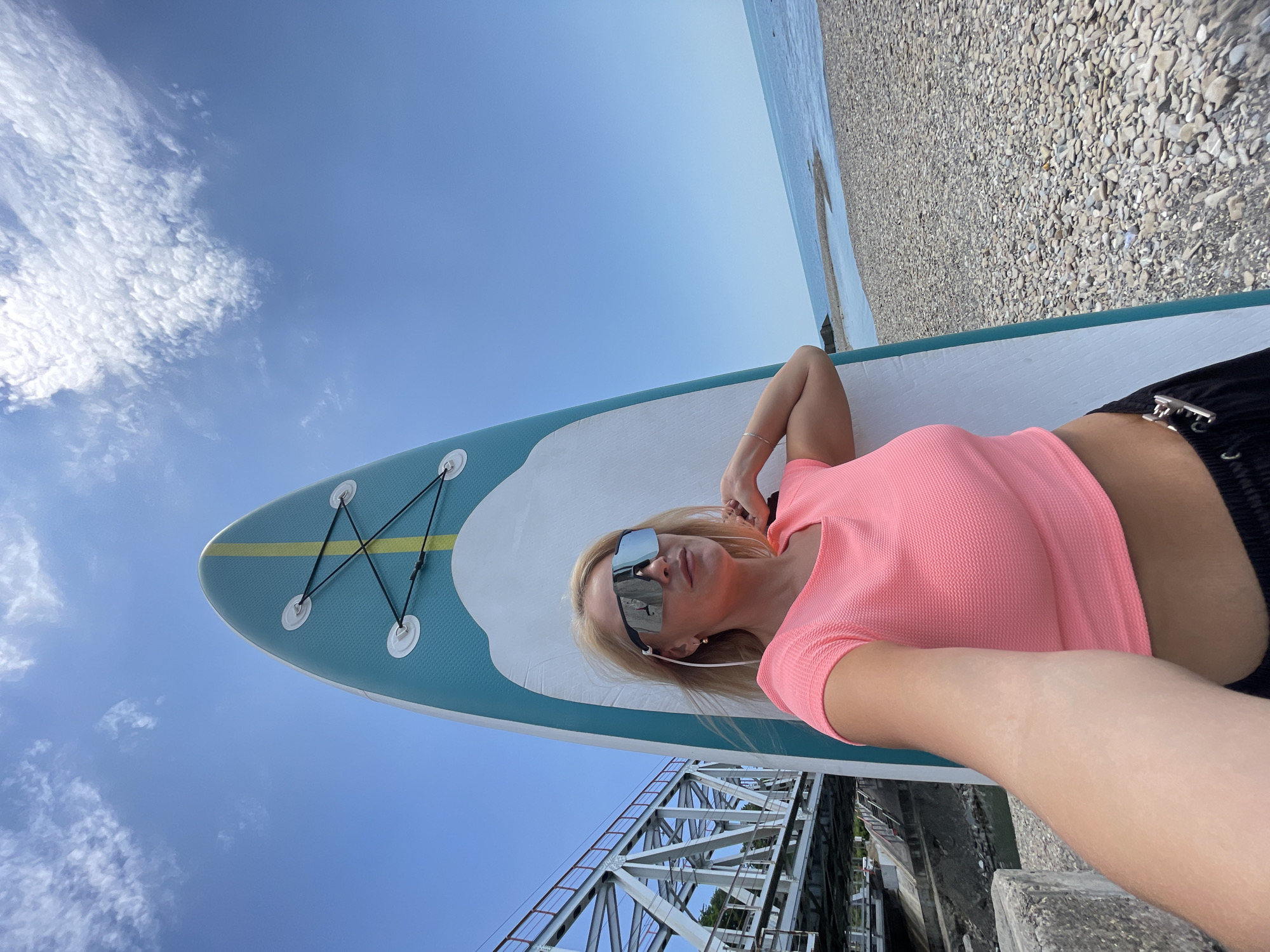 A blonde woman wearing sunglasses and a pink crop top poses for a selfie with a paddleboard. She is standing on a pebble beach next to the paddleboard, with a bridge and the ocean in the background. The sky is clear blue with some scattered clouds.