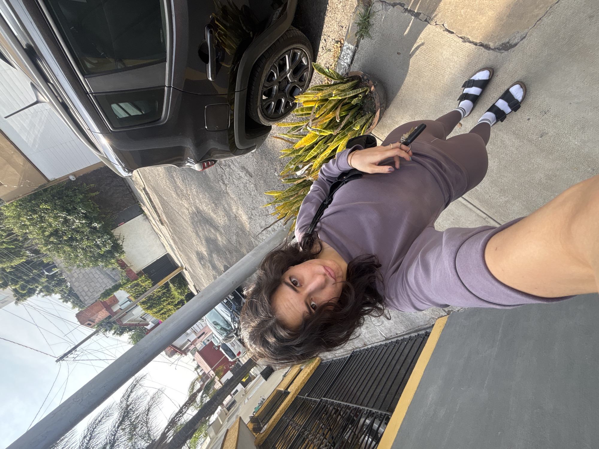 A selfie-style photo, rotated 90 degrees clockwise, captures a young woman standing outdoors on what appears to be a residential street in Puebla, Mexico. The woman, with dark, wavy shoulder-length hair, looks directly at the camera with a neutral expression. She is wearing a light purple long-sleeved top, dark plum or gray leggings, and black sandals paired with white crew socks. Keys are held in her left hand, which also shows rings on her fingers, and a black shoulder bag strap is visible across her body.
To her right, a black car, resembling an SUV or Jeep, is parked on the street. The car's black rims are visible, and the tire shows "BRIDGESTONE" branding with some tire specifications like "225/60R17" partially legible. Next to the car, a large potted plant, possibly a snake plant or Sansevieria, sits on the sidewalk. The concrete sidewalk and asphalt street are visible, indicating an urban or suburban setting.
The background reveals typical street scenes, including parts of other buildings, green foliage, and a prominent utility pole with numerous overhead wires stretching across the frame, a common sight in some Mexican cities. The sky appears clear and bright, suggesting a sunny day, likely morning or late afternoon, given the direction of the light and shadows. Another building with a yellow accent and dark security bars is visible behind the woman. The overall scene depicts a casual moment captured in an everyday outdoor environment.