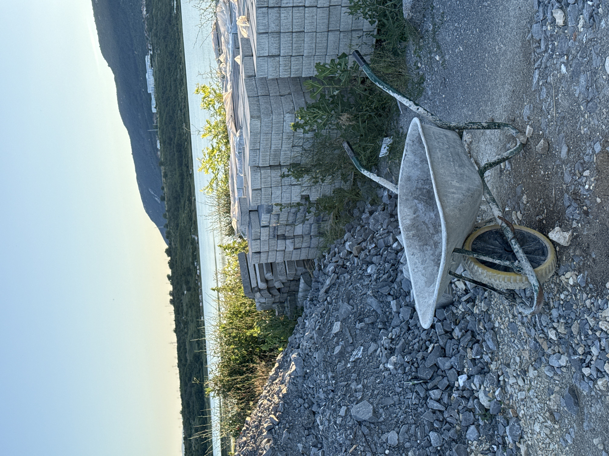 The image shows a wheelbarrow in the foreground on a gravelly surface. Behind the wheelbarrow, there is a large pile of grey stones, and further back, stacks of grey paving stones are visible, some covered with plastic sheeting. In the background, there is a body of water, with a tree-covered hillside and mountains beyond. The sky is clear and blue. The scene suggests a construction or landscaping site near a body of water, potentially in or near the city of Shumen, Bulgaria, though this specific location cannot be confirmed from the image alone.