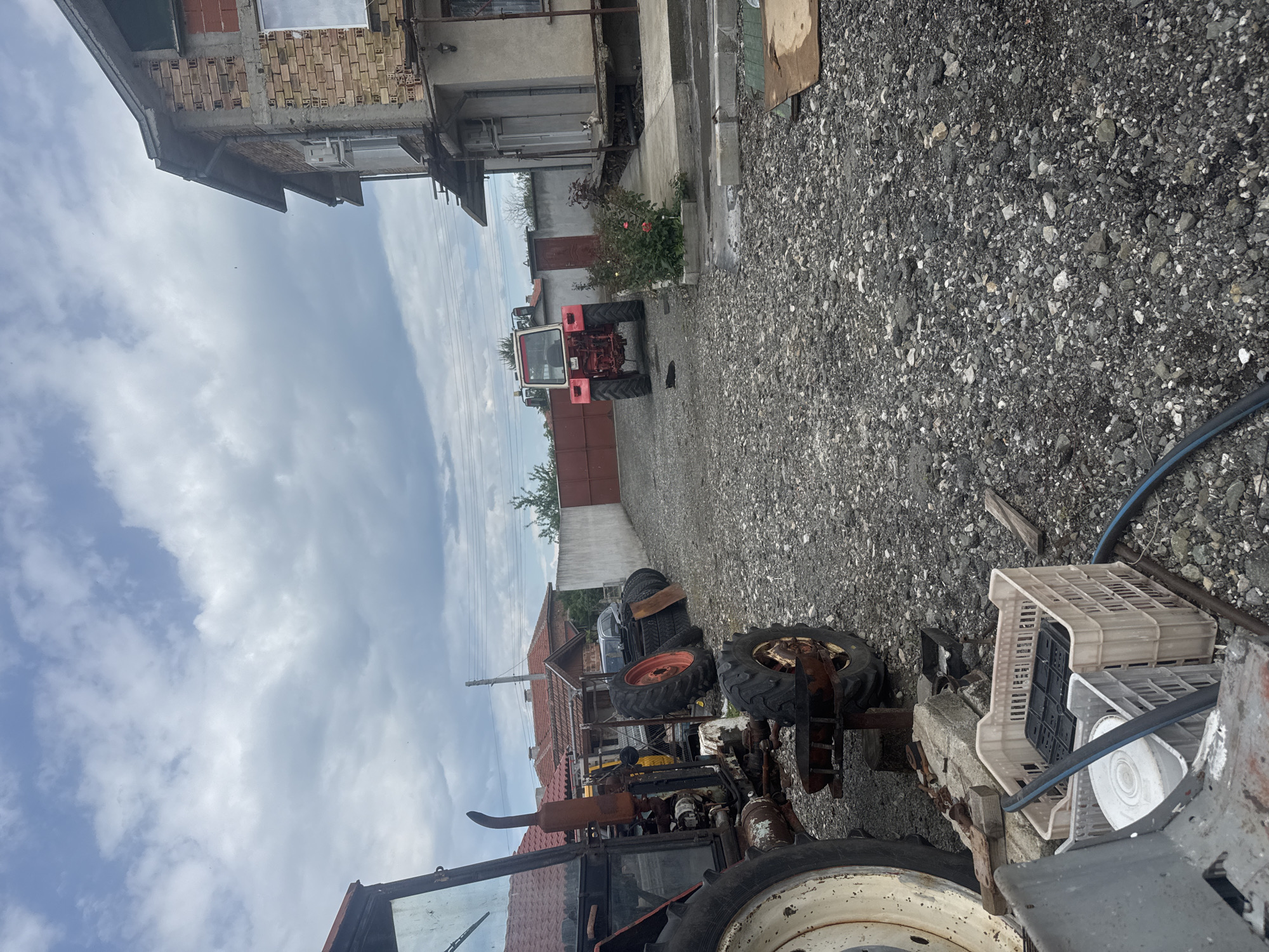 The image shows a yard in Sarnevo, Bulgaria. The ground is covered in gravel. In the foreground, there is a dilapidated tractor with missing parts and rust. Next to it are several plastic crates. Further back, tractor tires are stacked. A red and white tractor is parked in front of a building. The sky is overcast.