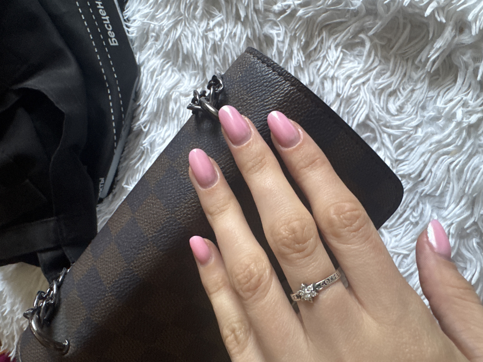 A right hand rests on a brown and black checkered clutch purse with a silver chain on a fuzzy white background. The hand features pink manicured nails and a silver ring with a diamond-like gem on the ring finger. A partially visible black bag with white lettering in Cyrillic script is located to the left.
