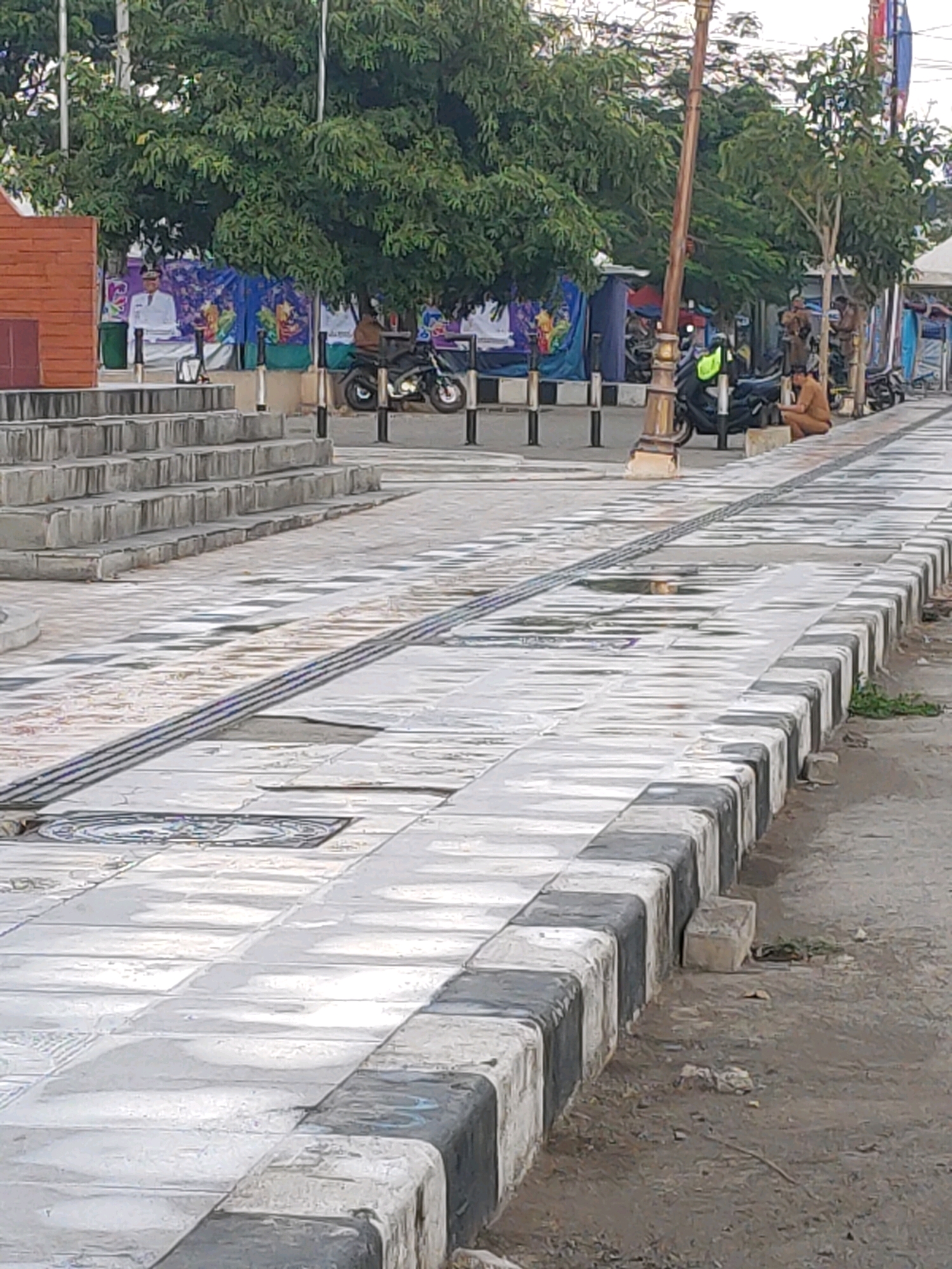 The image captures an outdoor scene in the city of Kanigoro, Indonesia. In the foreground, a paved walkway with a checkered pattern of light and dark tiles runs diagonally. The walkway is bordered by a curb with alternating black and white blocks. A drain runs along the center of the walkway.
In the middle ground, several people are visible. One person, dressed in brown, is sitting on the curb near a motorcycle. Other individuals appear to be standing or walking further down the path. A lamppost with a decorative base stands to the left of the walkway. Behind the walkway, a set of concrete stairs leads up to an area with trees and some temporary structures or booths with colorful banners. A person in a white uniform is standing near one of these structures. Motorcycles are parked in the area behind the walkway.
The background features lush green trees and what appears to be a bright sky, suggesting it is daytime. The overall environment seems to be a public space, possibly a park or a pedestrianized area, with elements of local culture evident in the banners and attire.