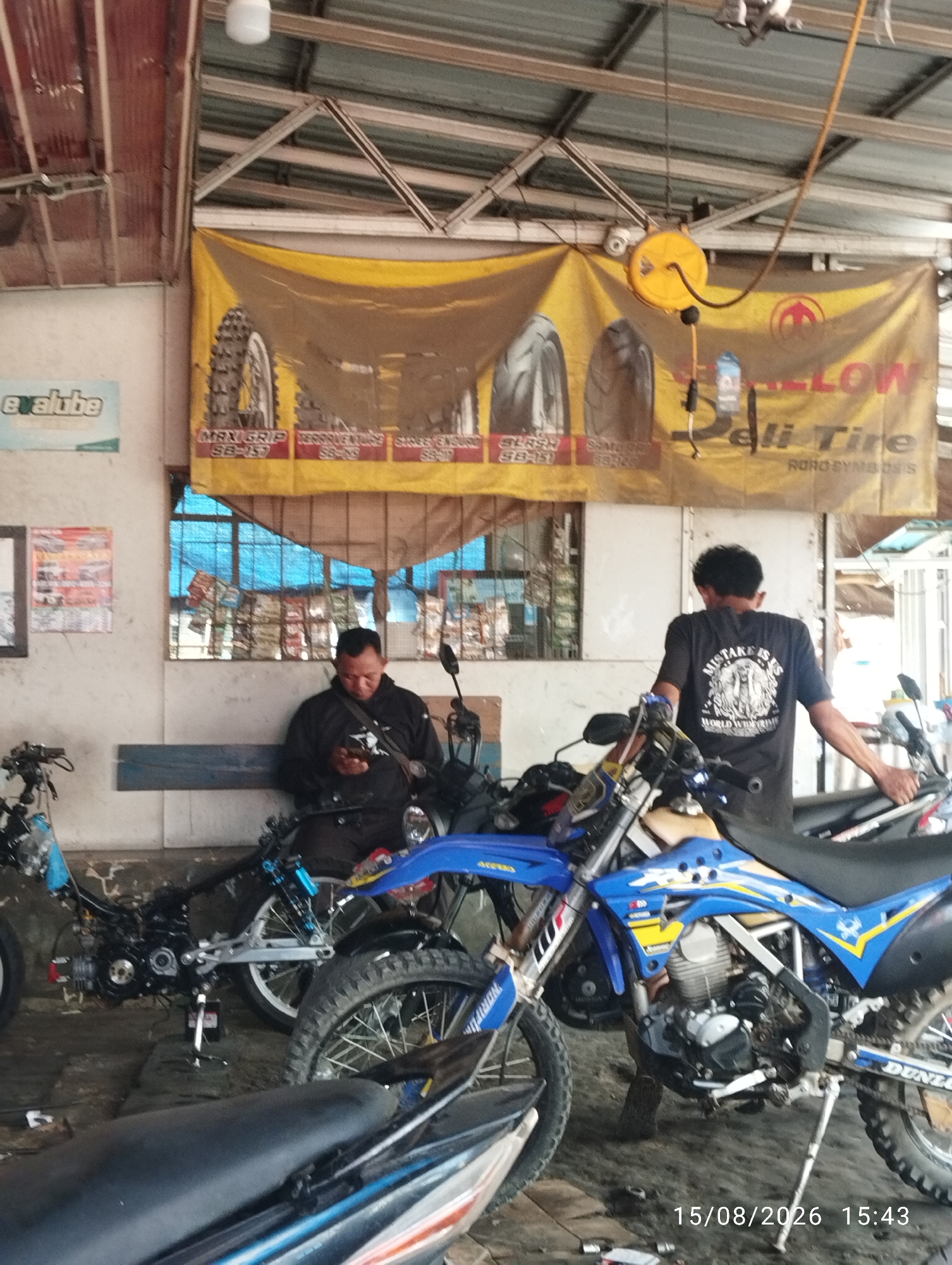 The image depicts a workshop scene in Tegal Rejo, Indonesia. Two men are present, one sitting on a bench and looking at his phone, while the other stands near a motorcycle. Several motorcycles, in various states of assembly and repair, are visible throughout the workshop. The workshop appears to be cluttered with tools and equipment, suggesting an active repair environment. A large yellow banner advertising tires hangs on the wall behind the men, featuring images of tire treads and brand names. The overall atmosphere is that of a busy motorcycle repair shop.
