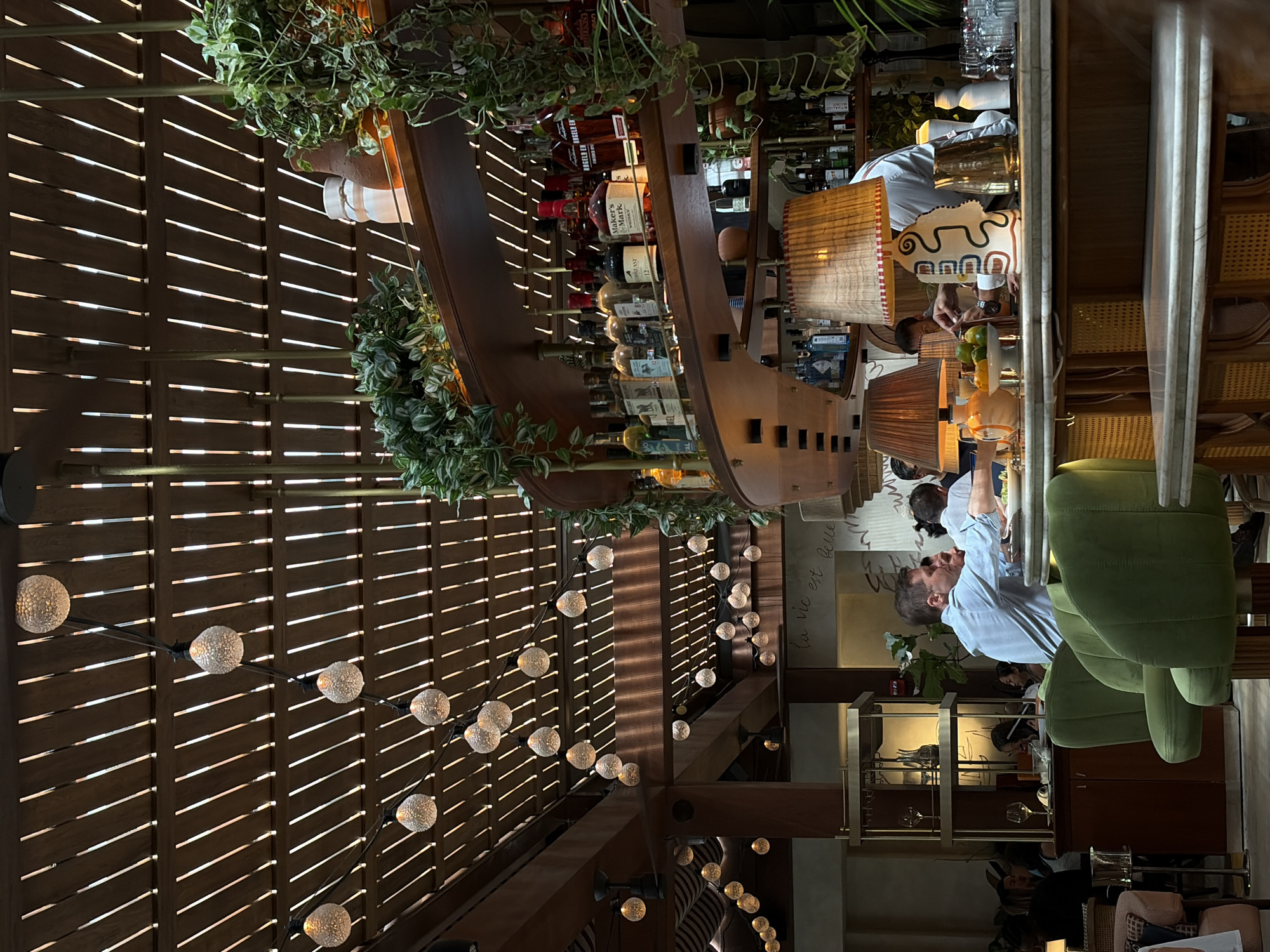 The image shows an interior scene of a restaurant. At the center is a curved bar with a wooden frame, stocked with bottles of liquor and adorned with greenery on top. Above the bar, a wooden lattice ceiling is visible, decorated with strings of globe lights. Patrons sit at the bar, interacting with the bartender. The furniture consists of green velvet chairs. In the background, there is a decorative shelf with glassware and artwork. The environment suggests a dimly lit, upscale establishment. The walls have a script with the words "La vie est belle". The location is in the city of Miami, United States of America.