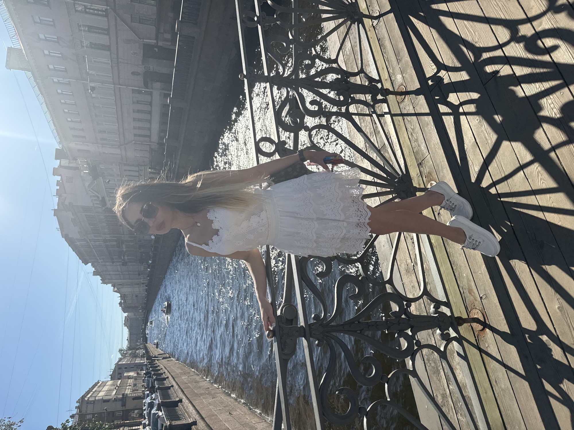 A young woman with blonde hair, wearing a white lace dress and white sneakers, stands on a wooden bridge with an ornate metal railing. She is looking down and to her right, with a slight smile. The bridge overlooks a canal lined with historic buildings, characteristic of Saint Petersburg, Russia. The water in the canal is choppy, reflecting the bright sunlight. Several small boats are visible on the water, and the sky is clear and blue. The time of day appears to be midday, indicated by the strong sunlight and distinct shadows cast by the bridge's railing onto the wooden planks. The overall mood is serene and picturesque. No visible text is present in the image.