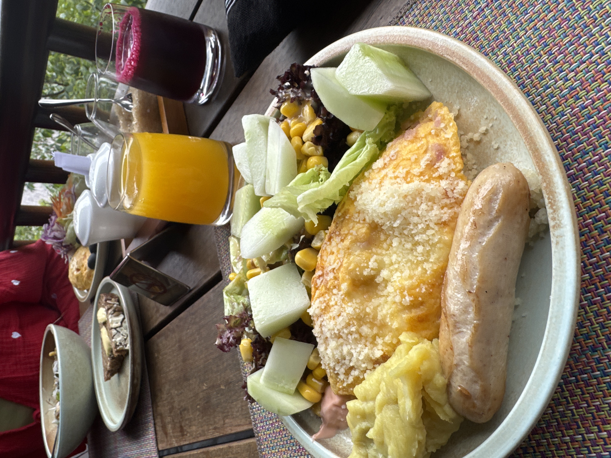 An outdoor breakfast or brunch table setting is depicted from a slightly elevated perspective. The primary focus is a light beige ceramic plate resting on a multi-colored woven placemat. On this plate, a golden-yellow omelet or frittata is prominently featured, heavily dusted with grated white cheese. Adjacent to it are a single light brown, cooked sausage, a scoop of pale yellow mashed potatoes or puree, and a small side salad. The salad comprises mixed green and red lettuce, sweet corn kernels, and several thick-cut pieces of light green cucumber or melon. A small dollop of pinkish sauce is visible near the mashed potatoes.
The rustic wooden slatted table holds additional items. To the left, a large light grey-green ceramic bowl containing an undiscernible dish is partially visible. Next to it, a small beige plate holds a slice of dark bread topped with a pat of butter. Further right, another small beige plate displays a light brown pastry or muffin accompanied by several black olives. Beverages include a tall glass of bright orange juice and a tall glass of dark red juice with a frothy surface. A wooden caddy centrally holds white ceramic sugar and condiment dispensers, a glass container with brown sugar and a spoon, and a small white holder with a plastic stirrer. A small brown, folded menu or place card stands upright on the table.
In the background, a person wearing a red garment with small white heart patterns is partially visible on the far left. Dark wooden railings delineate the edge of the dining area, beyond which a dense expanse of lush green trees and foliage indicates an outdoor setting in the natural environment of Púng Luông, Vietnam.