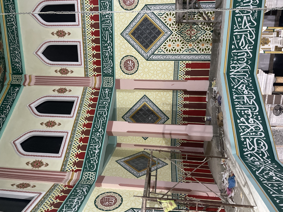 This image captures the richly decorated interior of a building, likely a mosque, in Bale, Indonesia, appearing to be in the final stages of renovation or construction. The walls and ceiling are adorned with elaborate Islamic art and calligraphy.
Dominating the scene are wide bands of green featuring intricate white Arabic calligraphy, which appear to run along the upper sections of the walls. Below these, sections of geometric patterns in red and brown hues add further detail. The main wall and ceiling surfaces are painted a light cream or beige, featuring subtle, repeating geometric patterns in a lighter tone.
Multiple arched windows with dark panes are visible along the upper walls. In the ceiling areas, diamond-shaped windows or vents with textured glass are set within intricate square borders of green, gold, and white. Circular medallions with Arabic script are also incorporated into the ceiling and wall designs.
The overall color palette is vibrant, comprising greens, reds, creams, gold, and light pink columns or supports. Metal scaffolding is present in the lower right and central parts of the image, alongside what appears to be construction debris, suggesting ongoing work. The intricate details and vibrant colors showcase a blend of traditional Islamic aesthetics and modern craftsmanship. The lighting suggests an indoor setting, possibly illuminated by artificial lights.