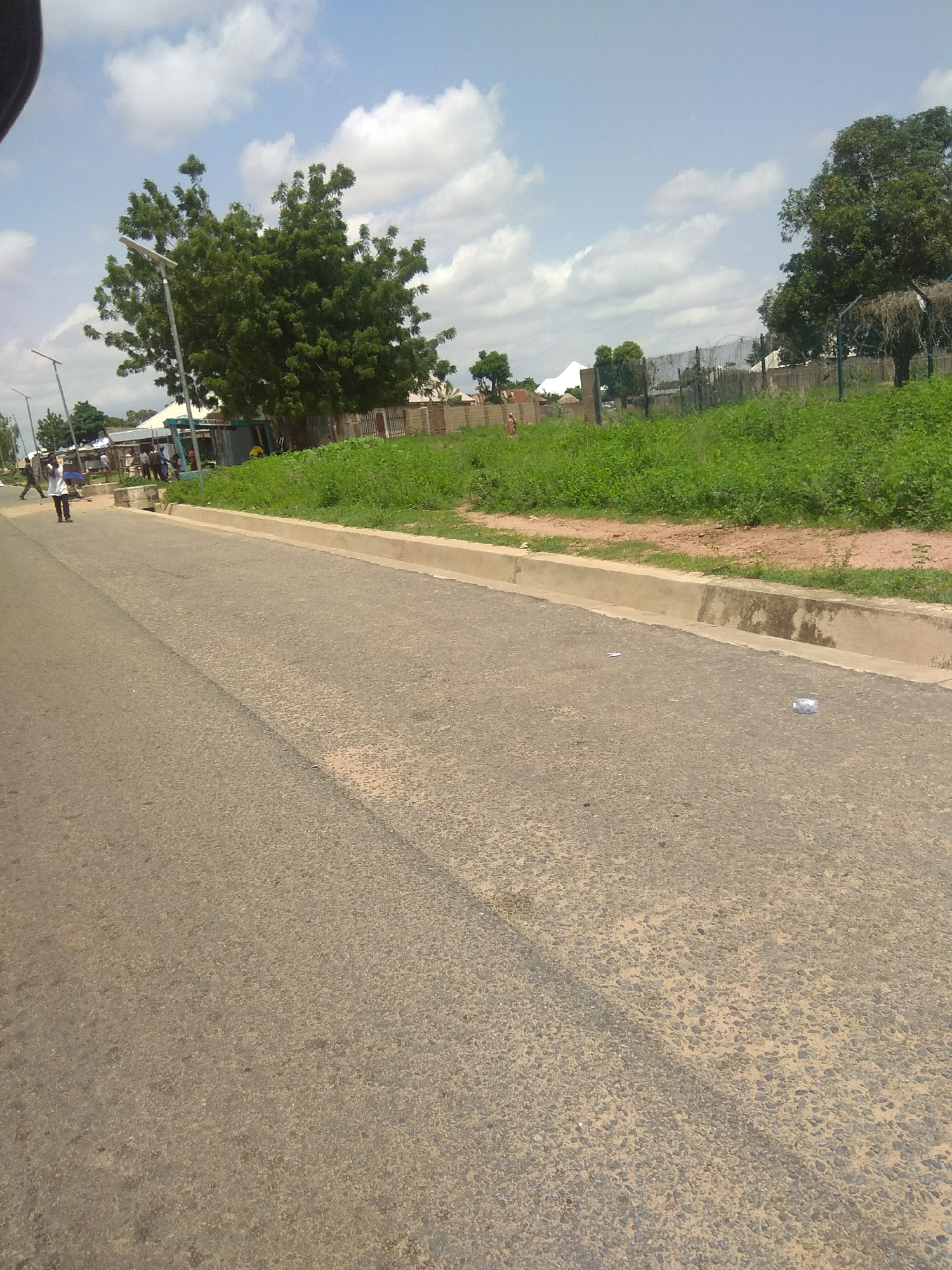 The image depicts a street scene in Jalingo, Nigeria, on a bright, cloudy day. A paved road occupies the foreground, with a distinct concrete curb separating it from a strip of green grass and vegetation. Beyond the grass, a low, brick wall with a wire fence runs horizontally across the midground. In the background, several trees with dense green foliage are visible, along with some buildings and a white tent-like structure. A few people are walking on the sidewalk or near the buildings. One person in the left midground is wearing a white top and dark pants, carrying a blue bag. Another person is further down the road. The sky is partly cloudy with white clouds against a pale blue background. The overall impression is of a typical urban streetscape in a sunny, tropical climate.