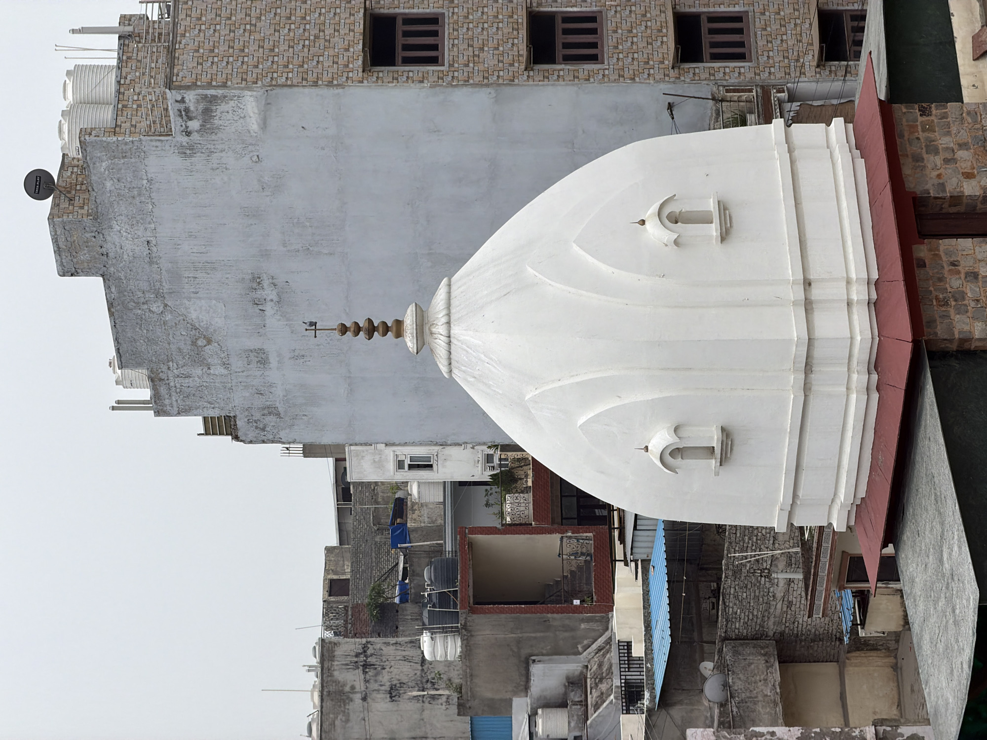 The image captures a rooftop view of buildings in a densely populated urban area, likely in Delhi, India, given the architectural style and the presence of water tanks on many rooftops, common in the region. Dominating the foreground is the white dome of a temple, a structure that appears to be relatively modern in its construction, characterized by its smooth, curved surface and a tiered base. Two small, arched niches are visible on the dome's façade, each containing a tiny finial. The dome is topped with a spire, adorned with multiple rounded sections and a final small spire.
Behind the temple dome, a multi-story building with a light grey, textured concrete facade stands tall. To its right, another building is clad in light brown and beige brickwork, featuring multiple floors with rows of dark-framed windows. Several water tanks, some white and some black, are visible on the rooftops of various structures scattered throughout the scene, indicating a reliance on stored water. The surrounding buildings are a mix of concrete, brick, and corrugated metal, with some showing signs of wear and tear. Blue tarpaulins are also visible on some rooftops, likely used for shelter or protection. The overall environment suggests a bustling city with a mixture of residential and possibly religious structures, viewed from an elevated perspective. There are no people visible in the image.