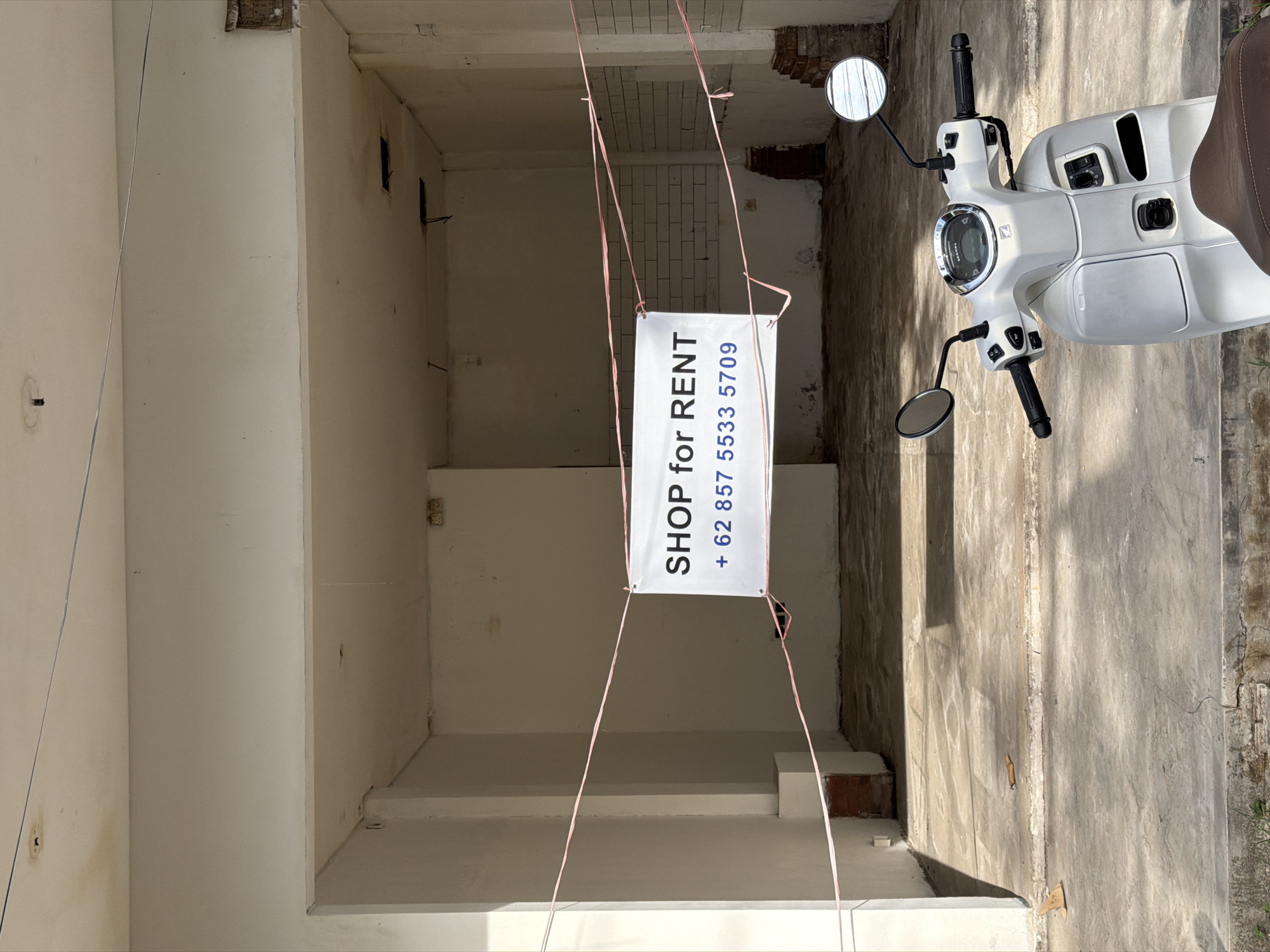The image depicts an empty shop space in Mengwi, Indonesia, indicated by a "SHOP for RENT" sign with the phone number +62 857 5533 5709. The shop appears to be undergoing renovation or is in a state of disrepair, with bare concrete walls and exposed ceilings. A white scooter is parked outside the shop, partially obscuring the entrance. The environment suggests a semi-urban or commercial area. No people are visible. The overall scene is of a vacant commercial space available for lease.