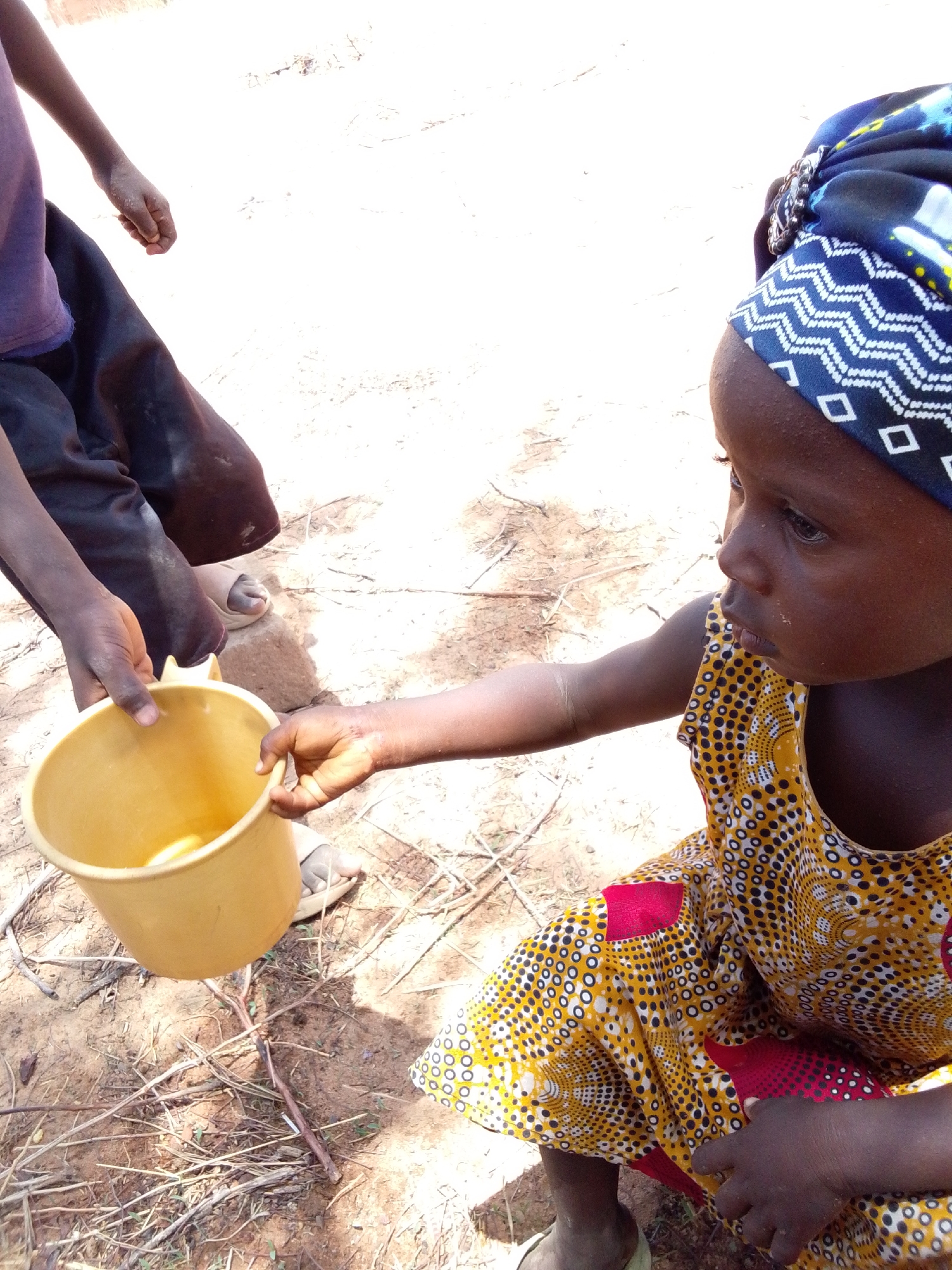 In Mulke Barikin Sayejo, Nigeria, a young girl wearing a patterned yellow and red dress and a blue patterned head covering is squatting. She extends her right hand towards a yellow plastic bucket being held by another individual whose legs and arm are partially visible. The ground is a mix of dirt, sand, and small twigs.