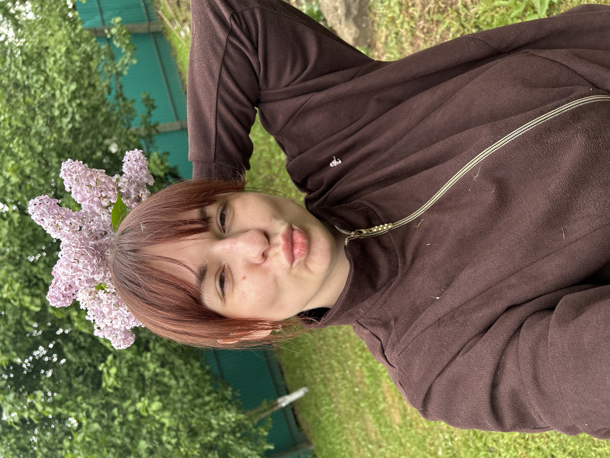A woman with reddish-brown hair is posing outdoors. She is wearing a brown, zippered sweatshirt. She has light pink flowers arranged around her head like a crown. The background includes green foliage, a green fence, and grass, suggesting a garden or park-like setting. The image is taken in Фоки, Russia.