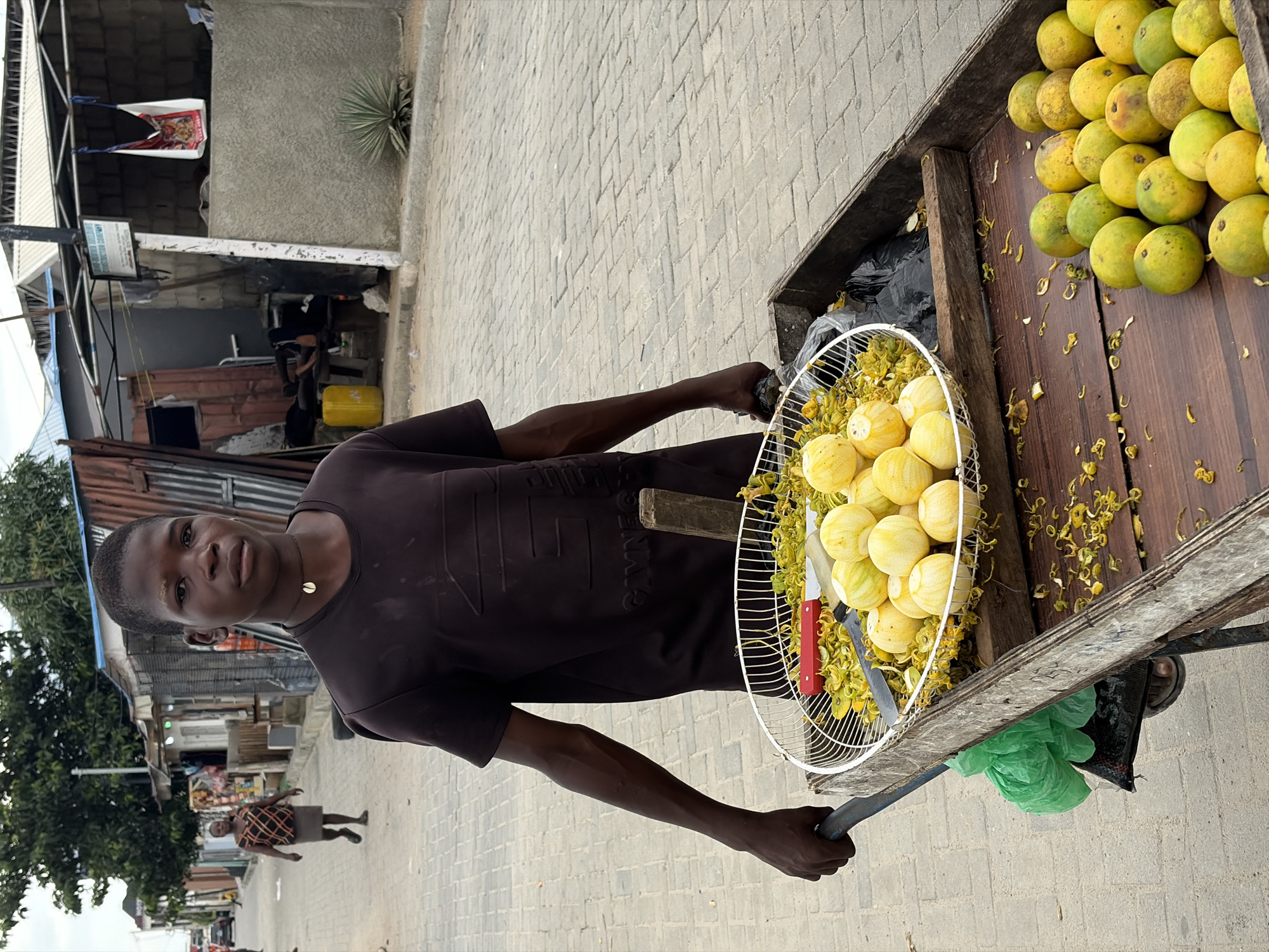 A young Black male vendor, appearing to be in his late teens or early twenties, stands in the foreground, pushing a wooden cart laden with fruits. He wears a dark brown t-shirt with embossed lettering and a simple seashell necklace. His gaze is directed towards the viewer.
The cart has two levels. The upper level contains a white wire basket filled with small, round, yellow fruits, possibly limes or lemons, and scattered yellow flower petals. A red-handled knife and a smaller knife are also visible in the basket. Below this basket, in a wooden compartment of the cart, a larger quantity of green and yellow fruits, similar in shape to oranges or tangerines, are arranged. A green plastic bag is attached to the side of the cart.
The scene is set on a paved street in Igboefon, Nigeria. The background reveals a street lined with various small shops and stalls, some constructed from corrugated metal and concrete. A woman in a patterned top and skirt walks away from the camera in the mid-ground, and other blurred figures and vehicles are visible further down the street. The environment suggests a bustling market or commercial area.