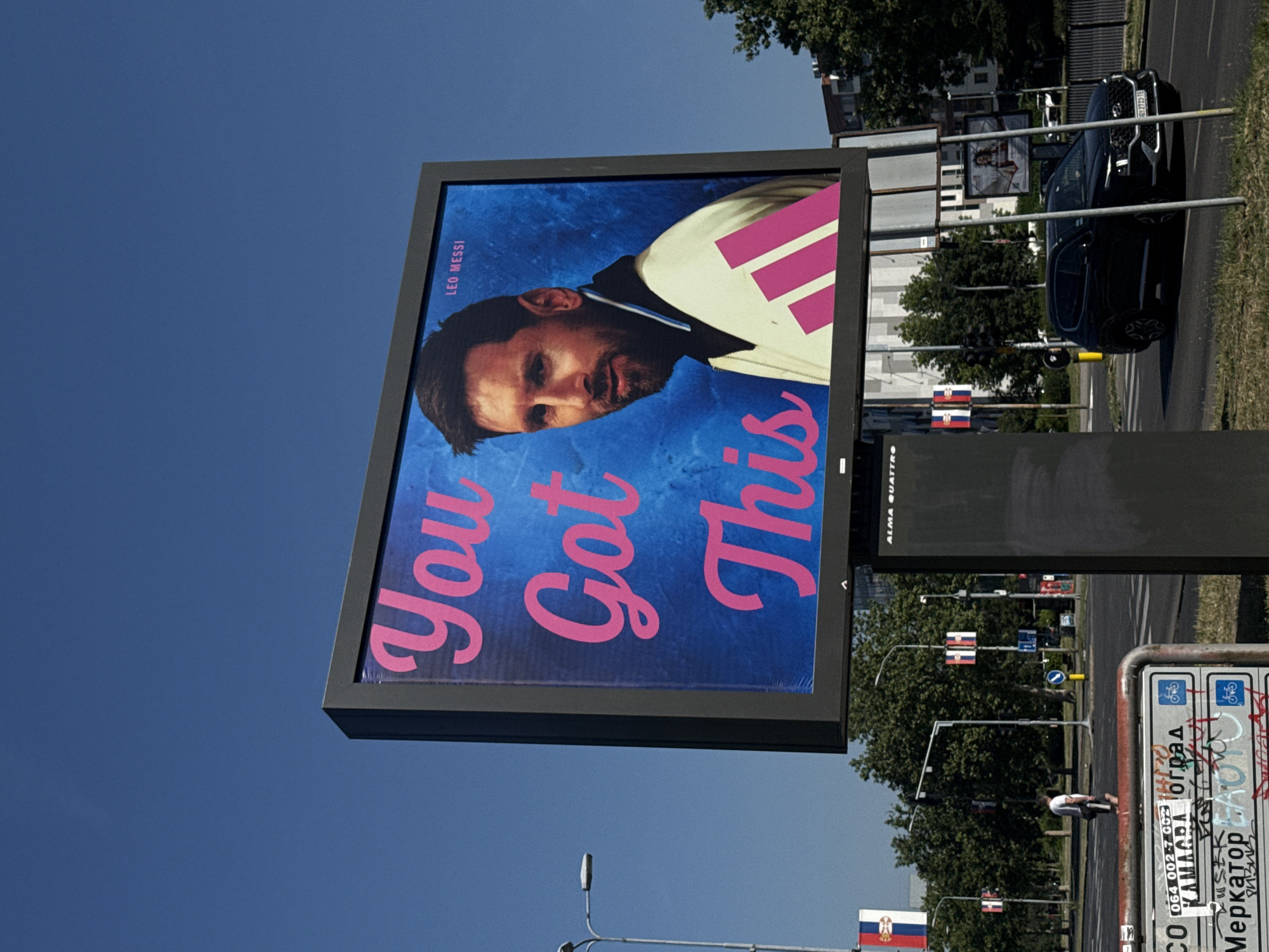 A large billboard displaying a portrait of Lionel Messi is visible against a clear blue sky. Messi is wearing a white jacket with pink accents and the text "You Got This" is superimposed in pink, stylized font. The billboard is situated in a city environment, likely Belgrade, Serbia, as indicated by Serbian flags and signage. The foreground shows a street with a black SUV driving on it, and in the immediate foreground, a metal railing with various signs, some covered in graffiti. Trees and buildings are visible in the background, suggesting an urban setting. The lighting indicates a daytime scene.