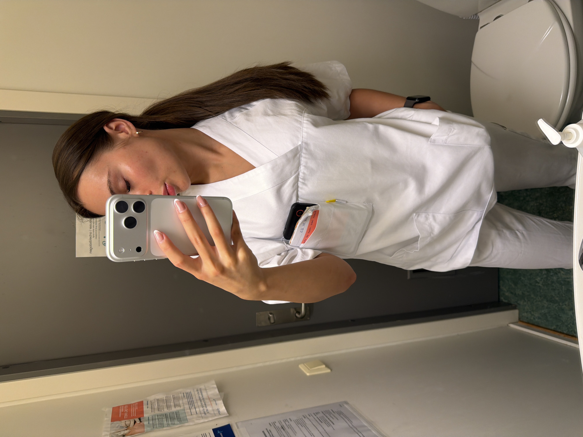 A young woman with dark brown hair pulled into a ponytail and wearing a white nurse's uniform takes a mirror selfie in what appears to be a restroom. She is holding a silver smartphone in her right hand, which is angled to capture her reflection. Her left hand is also visible, resting on her hip. A black smartwatch is on her left wrist. The uniform has a V-neck and pockets. In her left breast pocket, a black object, possibly a phone or card holder, and a yellow pen are visible. The background shows a gray door with a silver handle, a light-colored wall with some papers or documents attached, and a white toilet seat with a dispenser of hand soap. The flooring is a speckled green. The scene is well-lit. Based on the visible signs and the overall aesthetic, this location is consistent with a facility in Harstad, Norway, likely a hospital or clinic restroom.