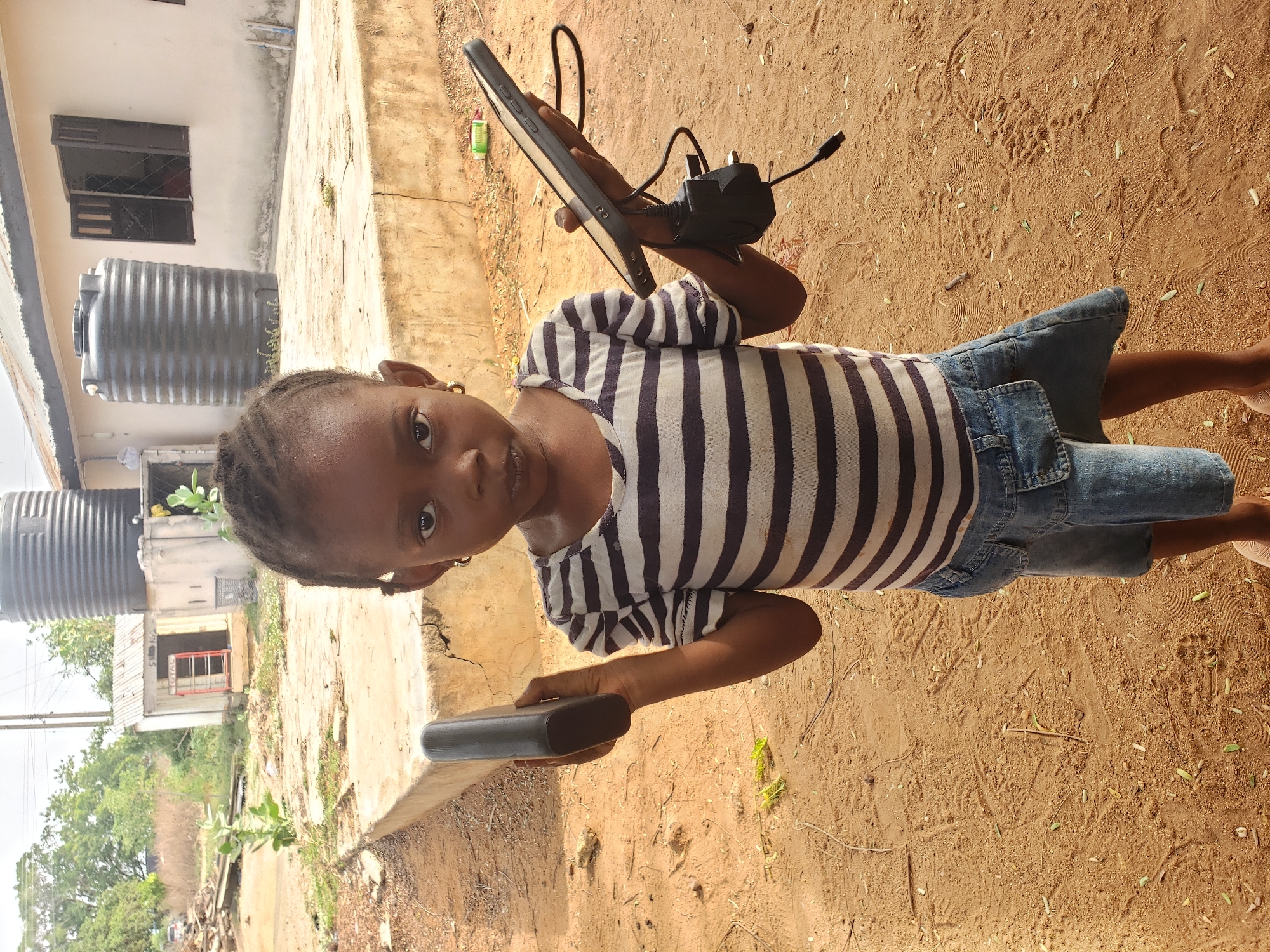 A young child stands outdoors in Adankolo, Nigeria, looking directly at the camera with a neutral expression. The child, likely a girl, has dark skin and braided hair, and is dressed in a white and dark striped short-sleeved top paired with denim shorts. In her left hand, she holds a black, rectangular device, possibly a power bank or a small electronic gadget. In her right hand, she holds a dark-colored tablet or a small laptop, which has a black power adapter with a UK-style plug and cable connected to it.
The setting appears to be a semi-urban or rural environment under bright daylight, indicated by clear shadows. The ground is dry, reddish-brown dirt, showing numerous footprints, small stones, and scattered dry leaves. A concrete curb or low wall runs alongside the dirt path. In the background, there is a light-colored building with an open, dark-framed window. Two large, black, ribbed water storage tanks are prominently situated next to this building. Further in the distance, another small structure with a red-framed opening is visible, on which some faint, blurry text resembling "TOTAL" can be seen. Green trees and foliage are present in the mid-ground and background, contributing to the outdoor atmosphere.