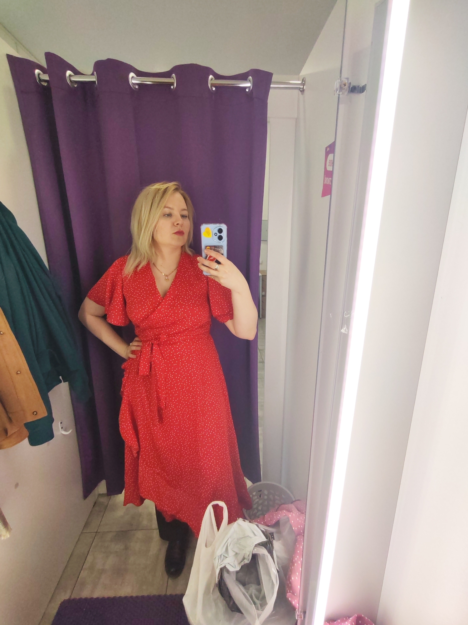 A blonde woman takes a mirror selfie in a retail store fitting room, located in Izhevsk, Russia. She is wearing a vibrant red wrap dress with white polka dots, featuring short butterfly sleeves and a tie-waist. Her hand rests on her hip, and she wears a gold necklace with a small pendant. Her phone, encased in a blue cover with a partial "Hello" inscription, is held up to capture her reflection. She is also wearing dark boots.
The fitting room features deep purple curtains with chrome grommets on the left, which likely serve as the entry. To the left of the woman, a dark green garment and a brown jacket hang on white hooks against a white wall. The floor is tiled, with a purple mat placed near the woman's feet.
The right side of the image shows a full-length mirror bordered by a bright vertical light fixture, illuminating the scene. In the reflection within the mirror, a white plastic shopping bag filled with items and a white laundry basket containing pink polka-dot fabric are visible on the floor. A price tag with "50%" clearly visible, along with some blurry Russian text (likely "Скидка!" for "Discount!"), hangs by a string near the mirror's edge, indicating a sale. The overall impression is that of a customer trying on clothes in a store.