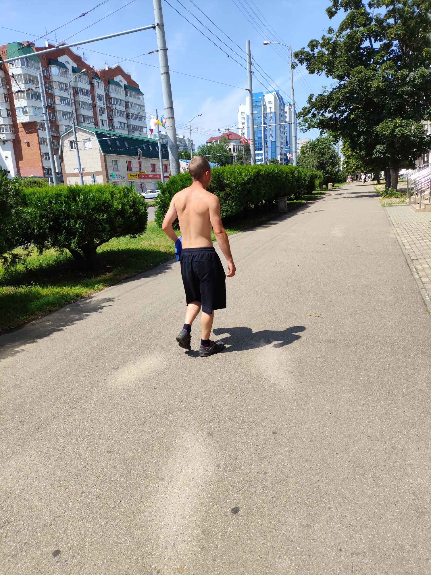 A shirtless man with short blonde hair is walking away from the camera on a paved path. He is wearing black shorts and dark athletic shoes with purple accents. The path is lined with green bushes on the left and a tree-lined sidewalk with people walking in the distance on the right. In the background, there are modern apartment buildings and a blue and white high-rise building. The image appears to have been taken in a city park or pedestrian area in Krasnodar, Russia, given the architecture and greenery. The sun is shining, casting shadows on the path.