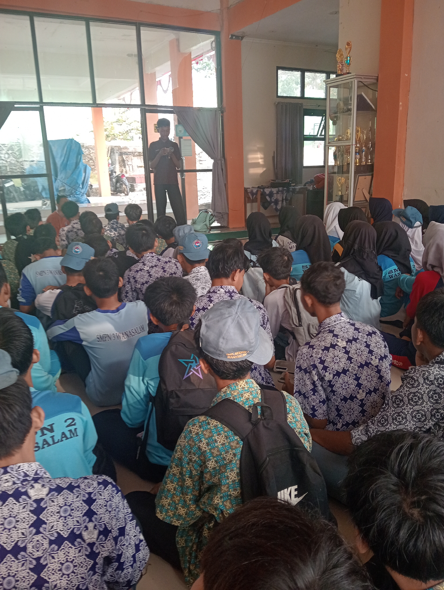 This is a medium shot photo taken inside what appears to be a school or community hall in Wanasalam, Indonesia, with a large glass-paned wall exposing an outdoor scene. The hall is filled with young students, likely a class or a school assembly, sitting on the floor. A young man stands by the glass wall, looking at his phone, dressed in a dark maroon shirt and black pants. In the background, through the glass, a motorcycle is visible outdoors. Inside the hall, a display cabinet filled with trophies stands to the right. Many students are wearing blue shirts with the text "SMP N 2 SALAM" or similar, and some have patterned batik shirts. The atmosphere seems to be one of a relaxed gathering or presentation.