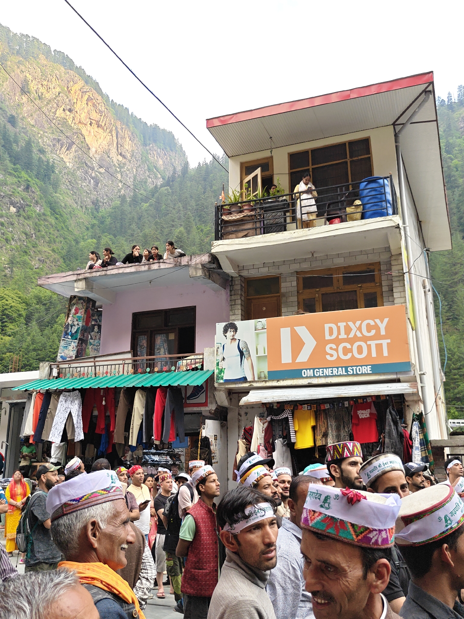 This image captures a bustling street scene in Manikaran, India, set against a backdrop of lush green, rocky mountains under a bright, possibly overcast, sky.
The scene is densely populated with people. On the street level, a large crowd, primarily men, is gathered. Many men wear traditional Himachali caps, some white with embroidered bands featuring religious or cultural text such as "महादेव ॐ" (Mahadev Om), "गढ़पति श्री ईबर महादा" (Garhpati Shri Ibar Mahada), "ॐ मैं गढ़पति" (Om Main Garhpati), and "कोठी पलाच" (Kothi Palach). Their expressions vary from engaged observation to smiles, suggesting a communal event or a lively market day.
People are also visible on the upper levels of the buildings. On a balcony of the central building, a woman stands near a blue water tank and potted plants. On the rooftop of the building to the left, several young women are seen looking down at the street.
The buildings feature shopfronts at the ground level. A prominent orange and blue sign reads "DIXCY SCOTT OM GENERAL STORE." Below it and to the left, racks display various garments, including pants and shirts, for sale. A red t-shirt with "DONALD DUCK" is clearly visible. The architecture is multi-story, with balconies and a mix of residential and commercial spaces. The overall impression is one of a vibrant community active during the daytime, surrounded by dramatic natural scenery.