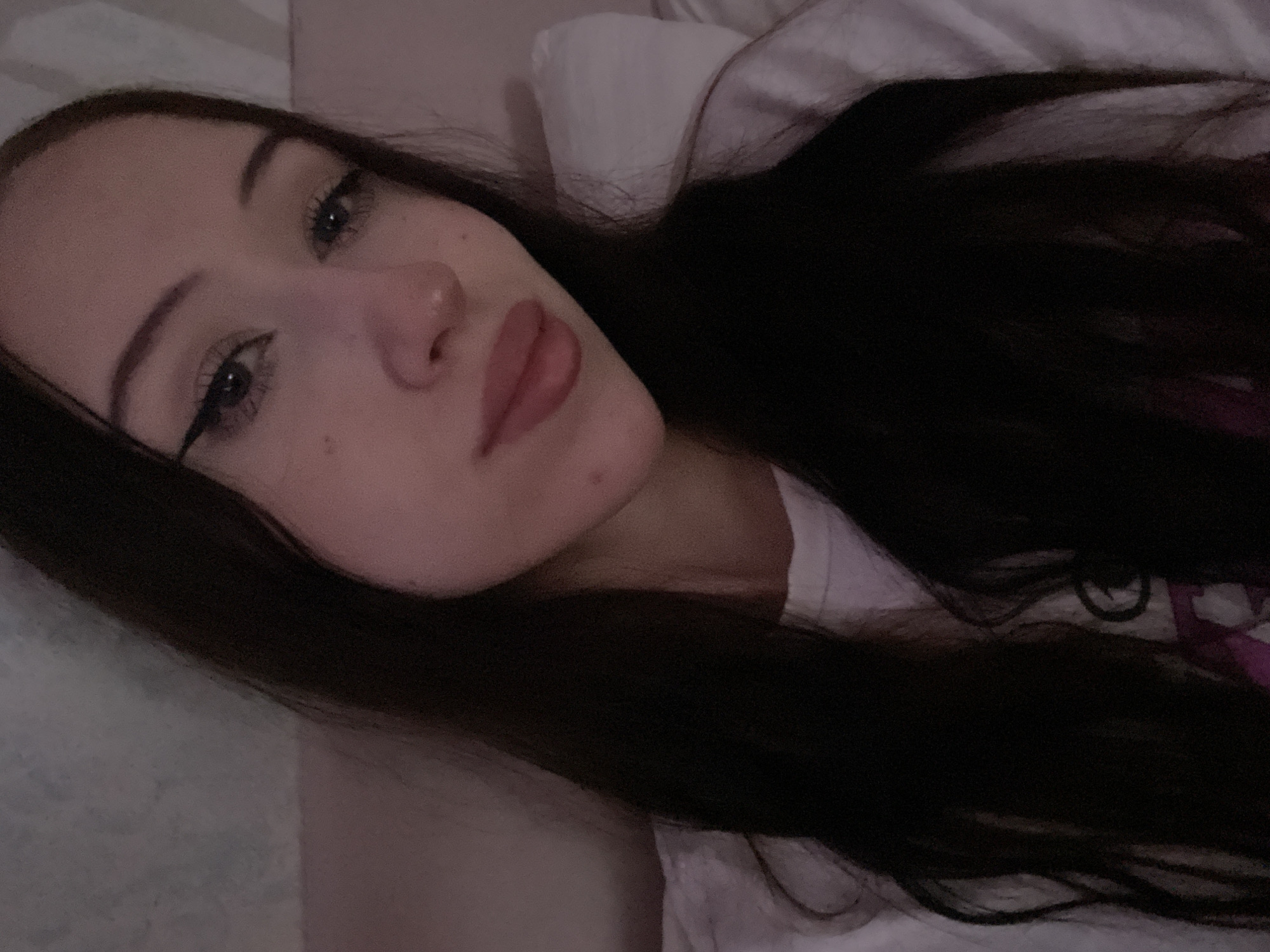 The media file is a close-up, low-angle selfie of a young woman lying down, with her head angled towards the viewer. She has long, dark hair that partially covers her face and shoulder. Her eyes are looking towards the camera, and she is wearing subtle eye makeup, including eyeliner. Her lips are slightly parted and appear to have a light pink lipstick on. The background suggests an indoor setting, possibly a bed, with soft, possibly wrinkled fabric that is a pale pinkish-white color. The lighting is dim, creating a soft, intimate mood. There is no visible text in the image. The overall impression is a candid, relaxed moment captured in a private setting.