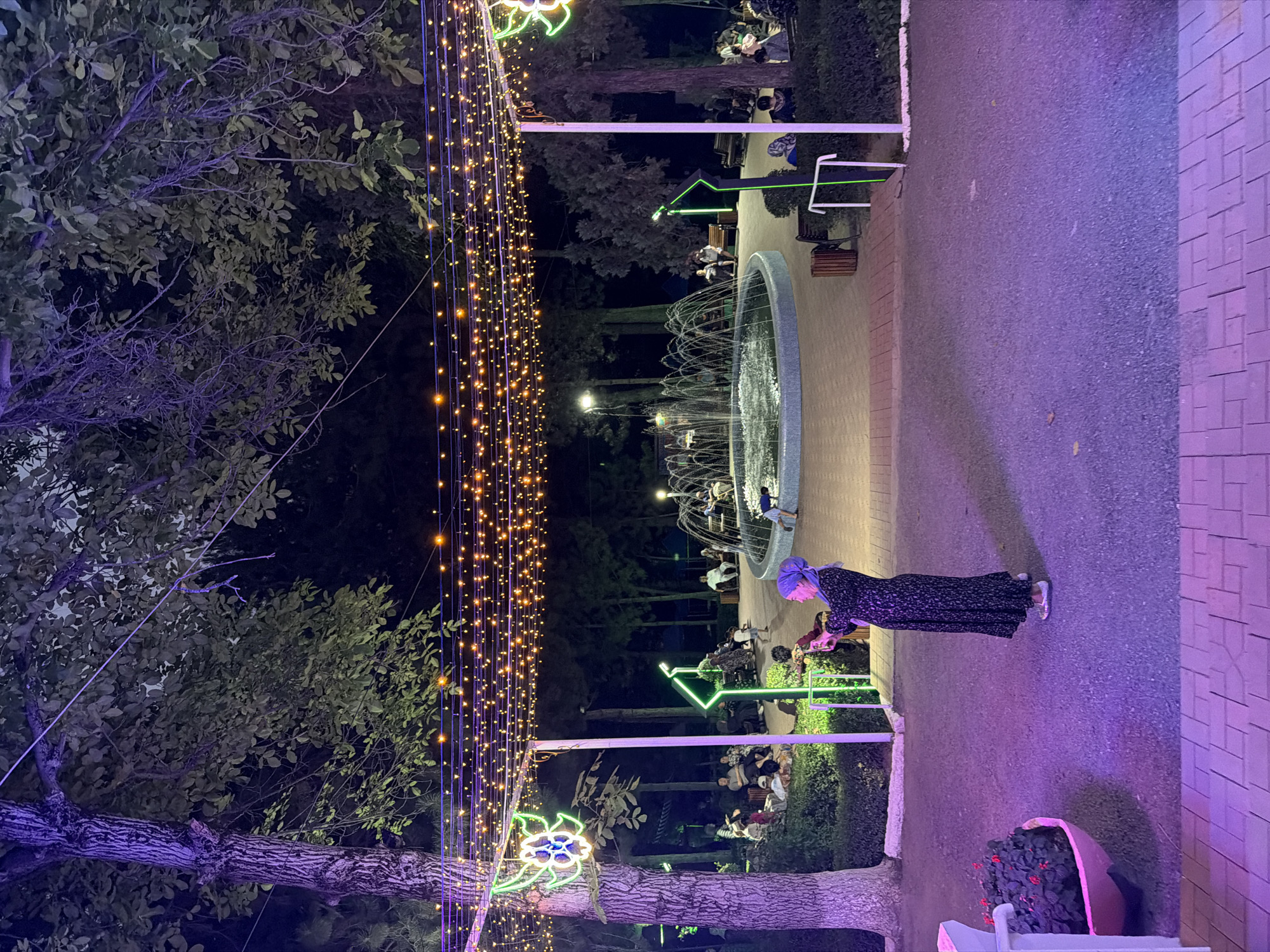 An outdoor evening scene in the city of Khumsan, Uzbekistan features a woman in a long, dark floral dress and a light purple headscarf standing on a paved walkway. She is holding a smartphone in her hands, appearing to take a photo or video. The walkway is lit with purple lights from below and surrounded by dark gravel. To the left, a planter filled with dark green foliage and red flowers is partially visible, also illuminated by purple light. Beyond the walkway, a park area is visible with trees, benches, and a central fountain. The park is adorned with numerous strings of small, warm-toned lights overhead, creating a festive atmosphere. Several groups of people are seated and standing around the fountain and on benches, enjoying the evening. The trees are dark and the sky is black, indicating it is nighttime. The overall environment is an outdoor public space, likely a park or plaza, decorated with lights for an event or regular evening illumination.